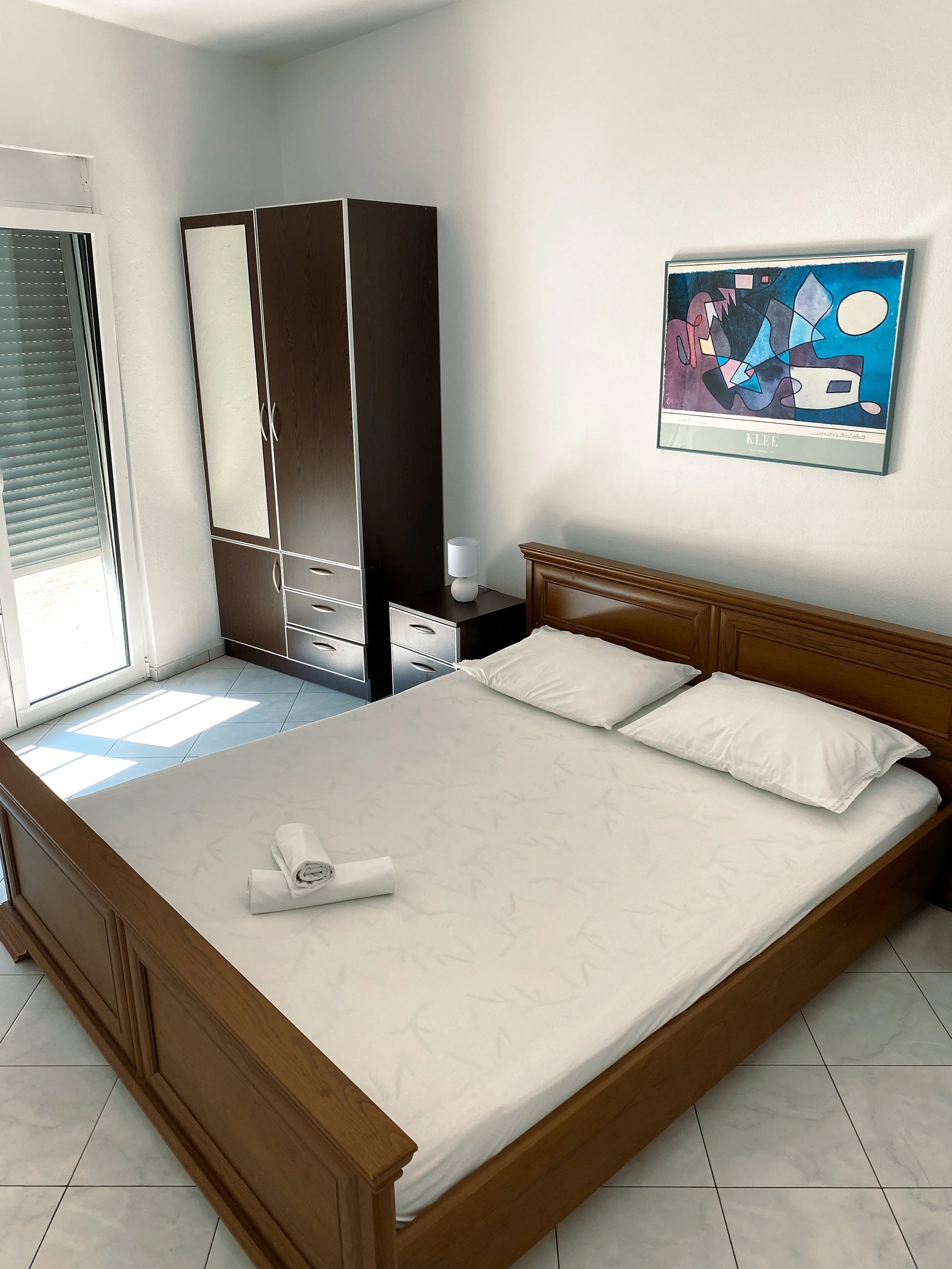 A neatly made bed with white sheets and pillows, a rolled towel, wooden headboard, nightstand with lamp, dark brown wardrobe, and a colorful abstract painting on the wall in a bright room with sliding glass door.