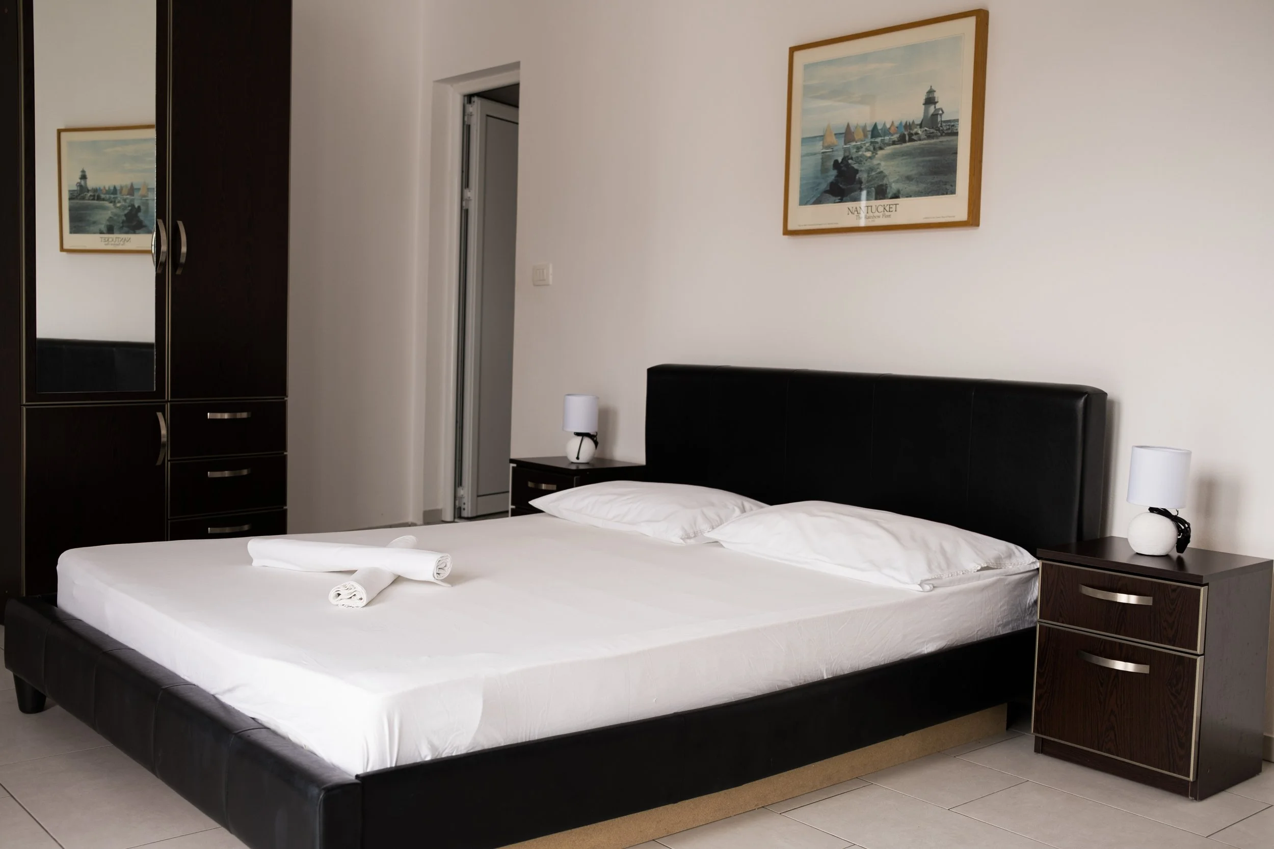 A neatly made bed with white linens and pillow, with two rolled towels on top, in a hotel room. There are two bedside tables with white lamps, a framed nautical artwork on the wall, a black wardrobe with mirrors, and a door leading to a small closet 
