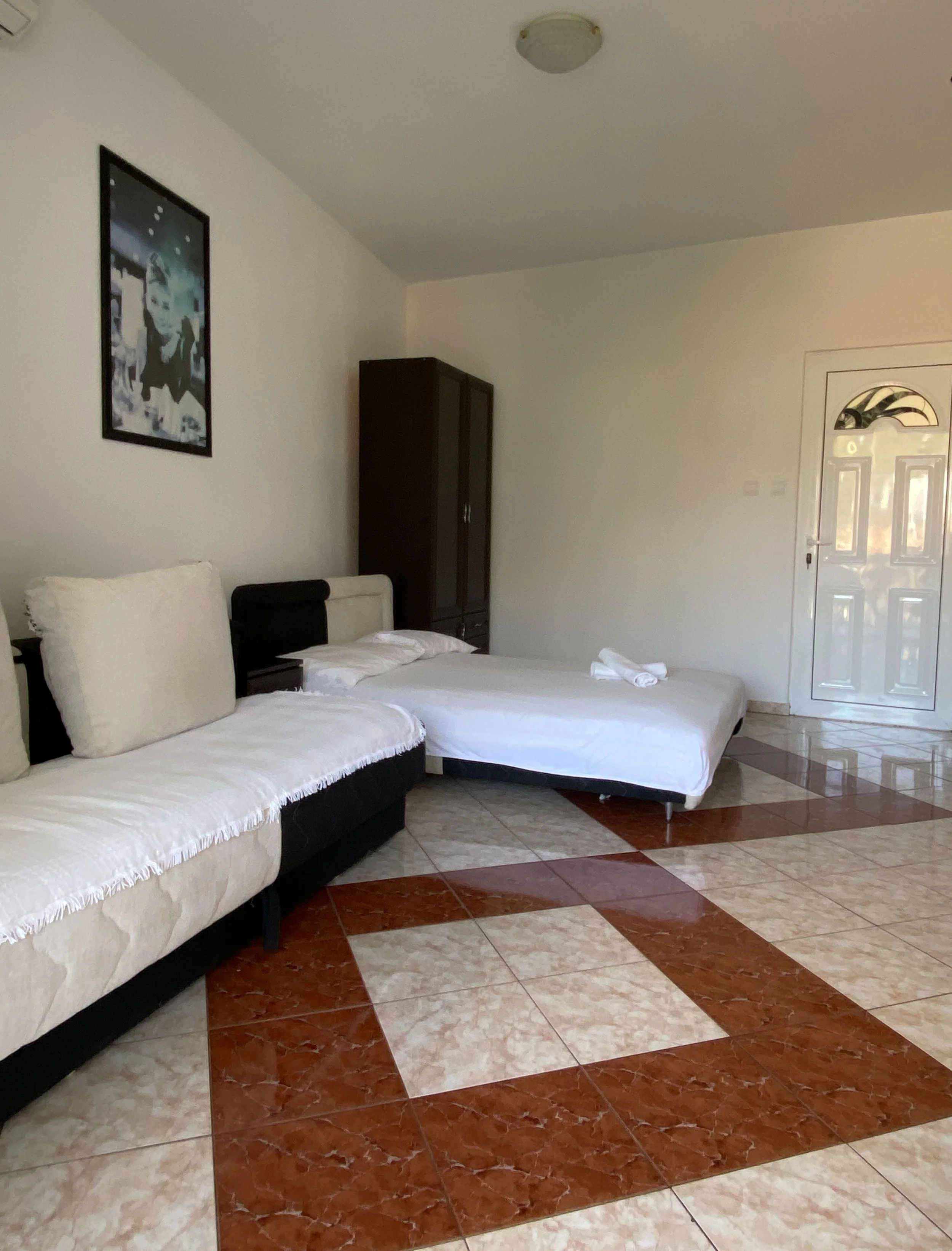 A hotel room with a white and beige sofa, a bed with white linens, a dark wooden wardrobe, a door, and a colorful tiled floor.