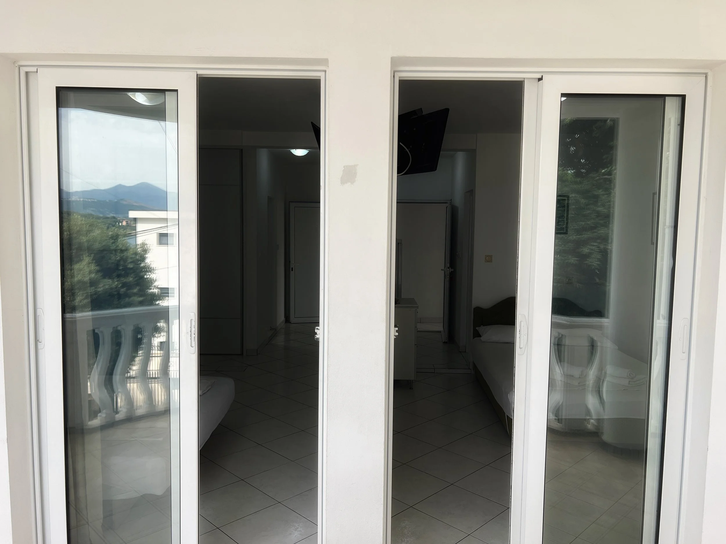 View through a glass sliding door showing reflections of mountains, trees, and a building outside, with an interior room containing a bed and a TV visible in the background.