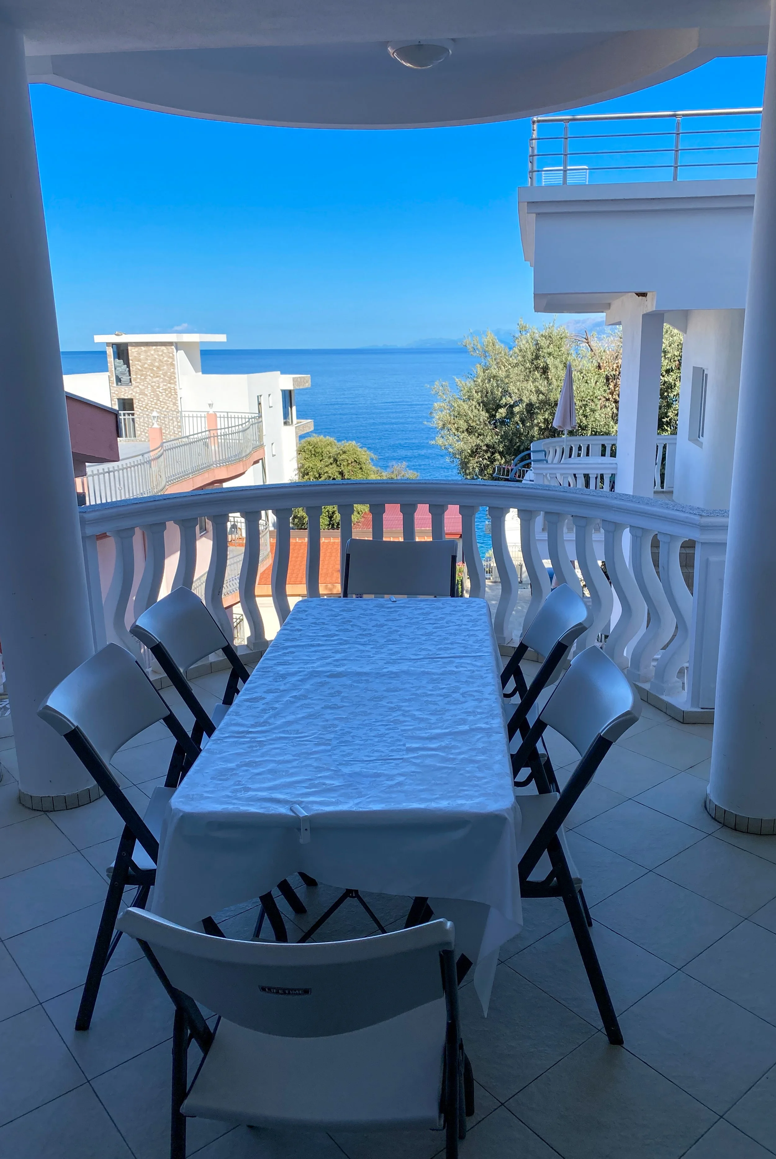 Balcony with a table covered by a white tablecloth, surrounded by six chairs, overlooking the ocean with blue sky and nearby buildings.