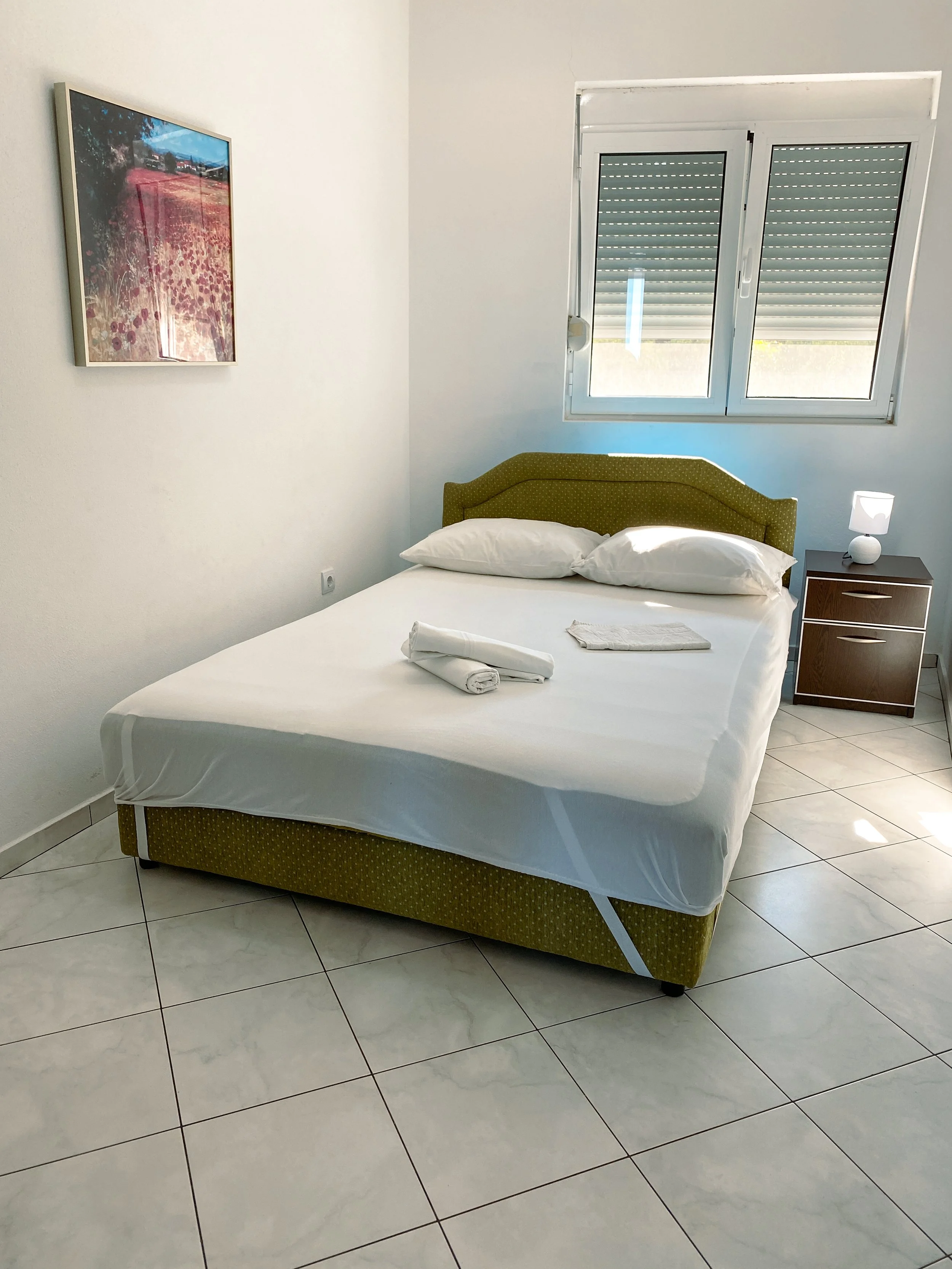 Hotel room with a bed, two pillows, a nightstand with a small lamp, a window with blinds, a towel, and a wall painting.