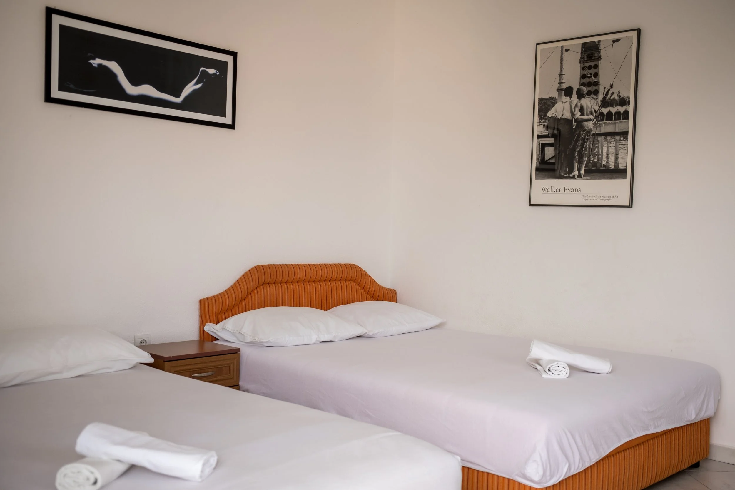 Two beds in a hotel room with white bedding, an orange headboard, and two rolled towels on each bed. Black and white photographs on the wall, one of a woman and one titled Walker Evans, are hung above the beds.