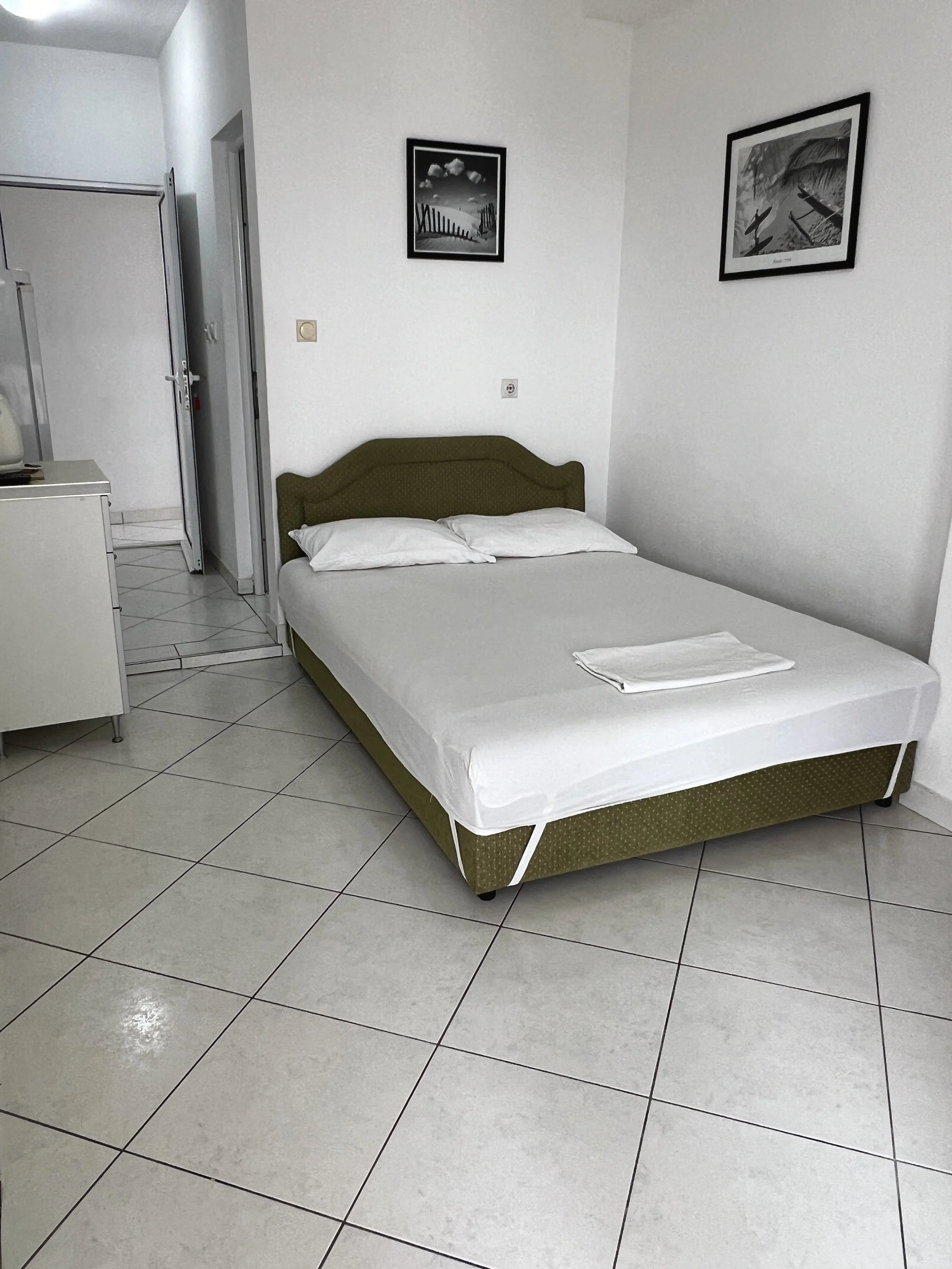 A hotel room features a bed with white sheets and pillowcases, a green upholstered headboard, two pillows, and a folded towel on the bed. The room has white tiled flooring, white walls, and two black-and-white framed pictures hanging above the bed. T