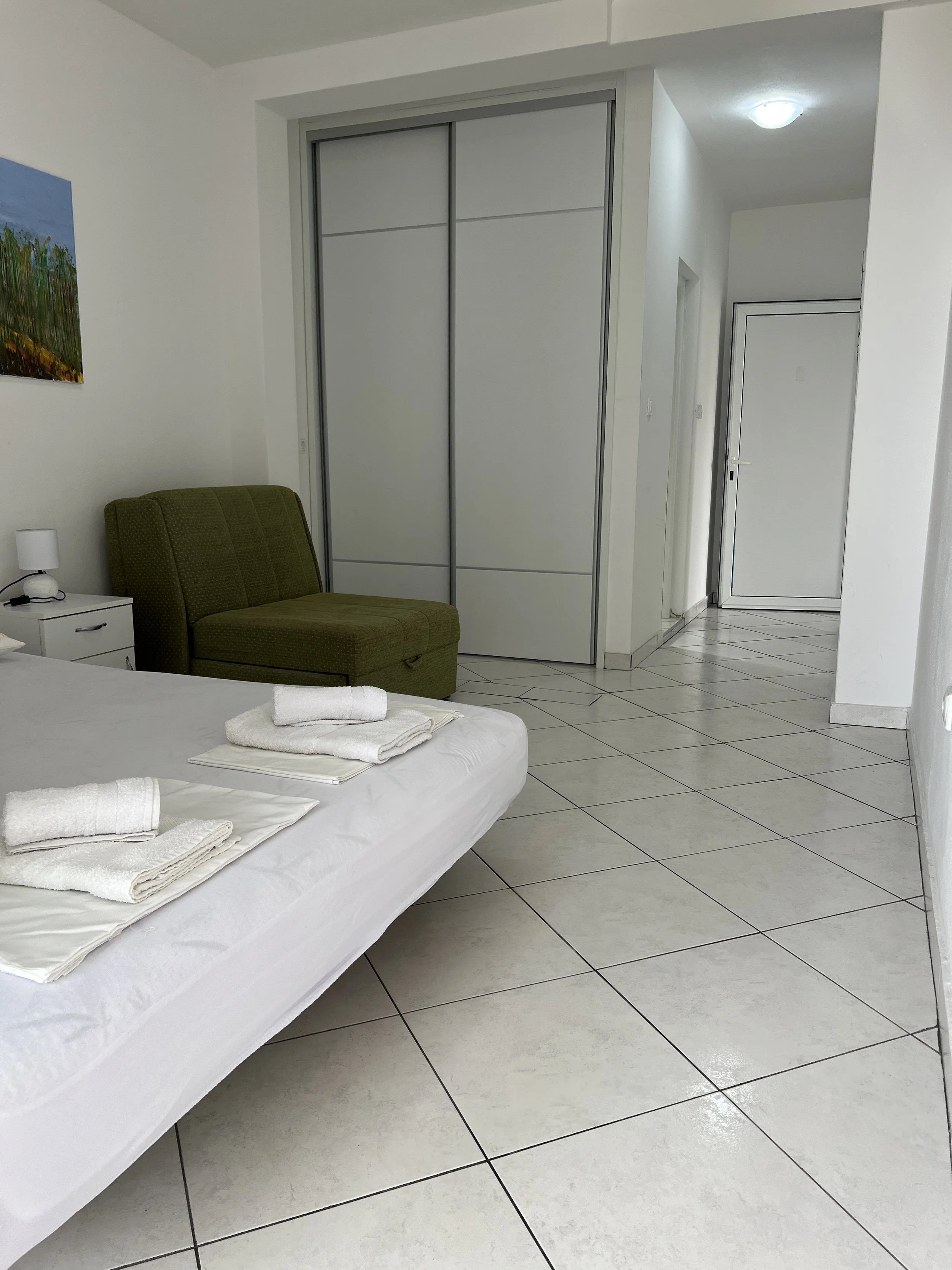 A hotel room with a white bed neatly made, with three folded towels on top, next to a white nightstand with a lamp. A green upholstered armchair is positioned next to a closet with closed sliding doors. The room has white tiled floors and white walls