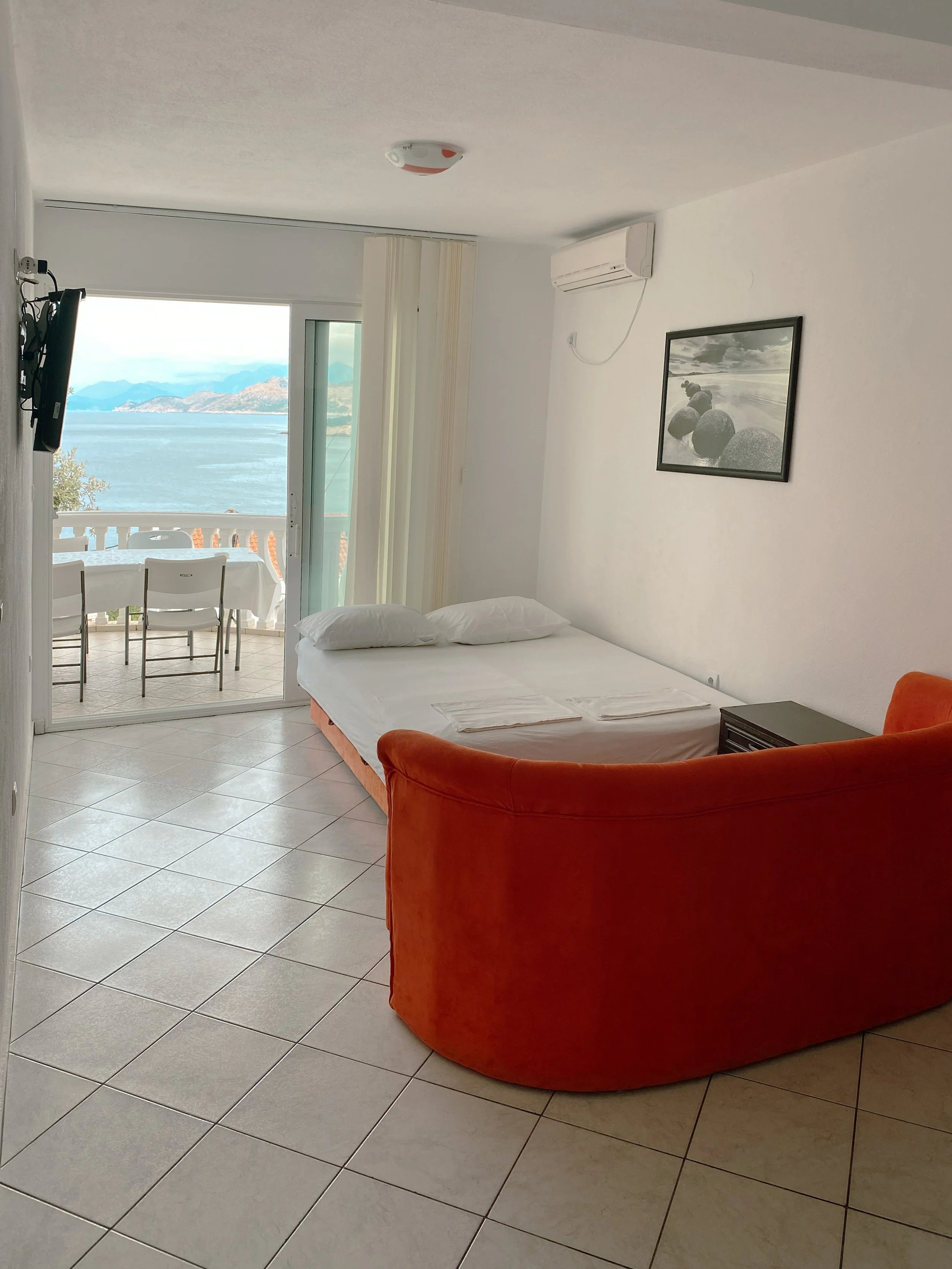 A hotel room with a balcony view of the water and mountains, a double bed with white linens, black nightstand, orange sofa, wall-mounted TV, black and white framed photo, air conditioning unit, and sliding glass door leading to the balcony with outdo