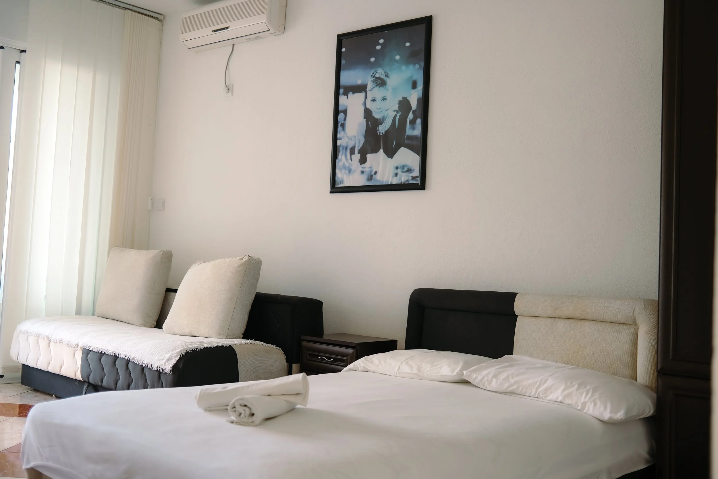 A bedroom with a bed, pillows, and a rolled towel, a black and white photograph on the wall, a sofa with cushions, an air conditioning unit, and window curtains.