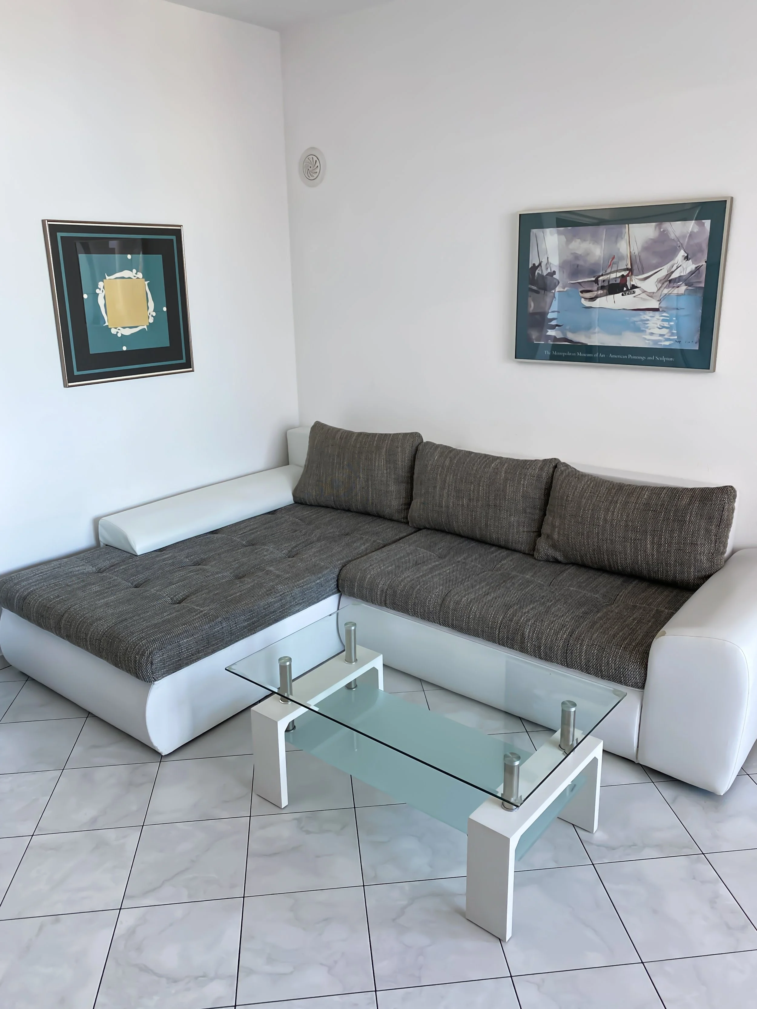 Living room with a white and gray sectional sofa, two framed paintings on white walls, and a glass coffee table on white tiled floor.