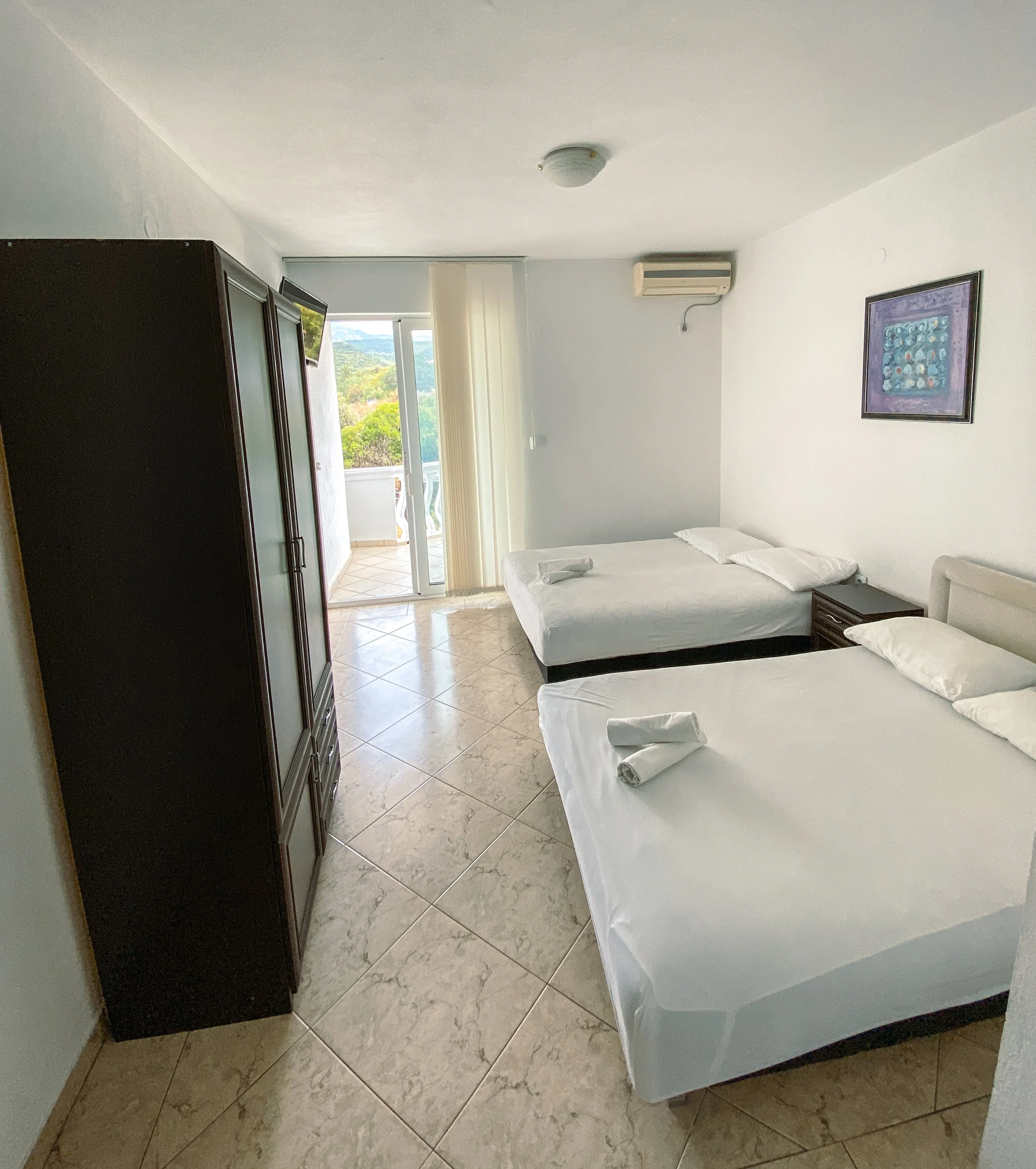 Hotel room with two beds, a wardrobe, a nightstand, a sliding glass door leading to a balcony, and a framed painting on the wall.