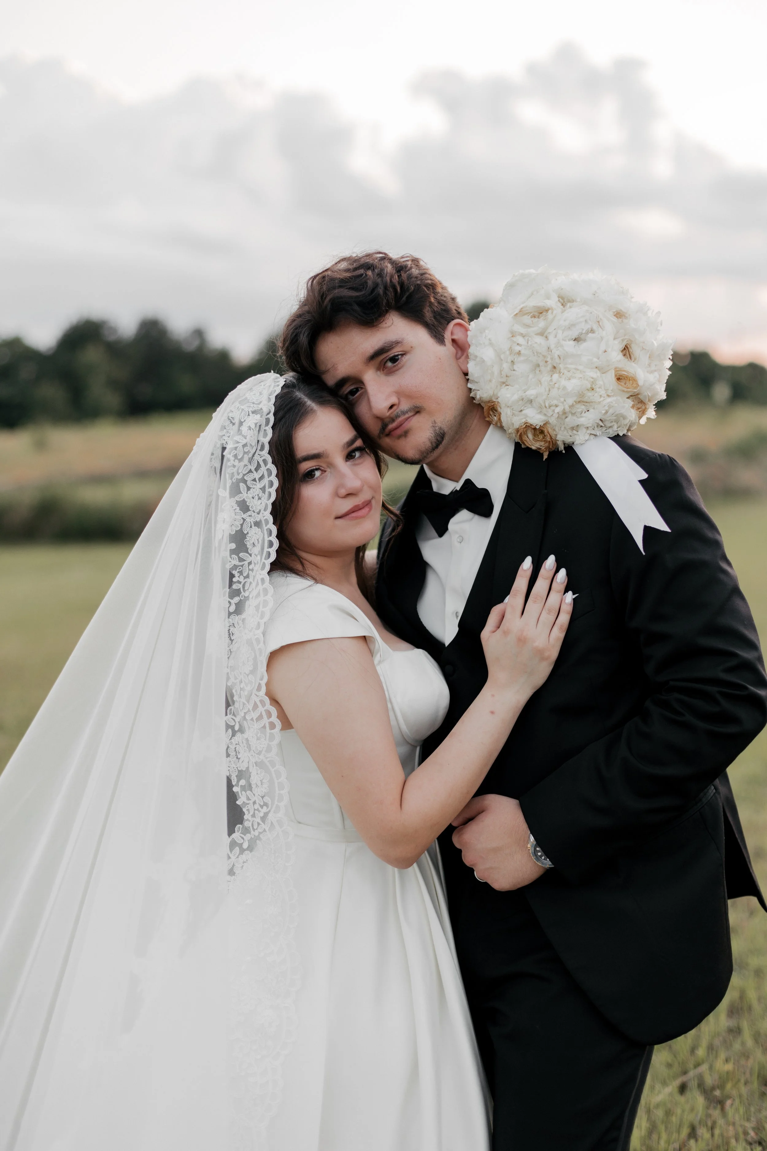 A bride and groom in wedding attire posing outdoors. The bride wears a white dress and lace veil, and the groom wears a black tuxedo with a bow tie. They embrace each other with a field and cloudy sky in the background.
