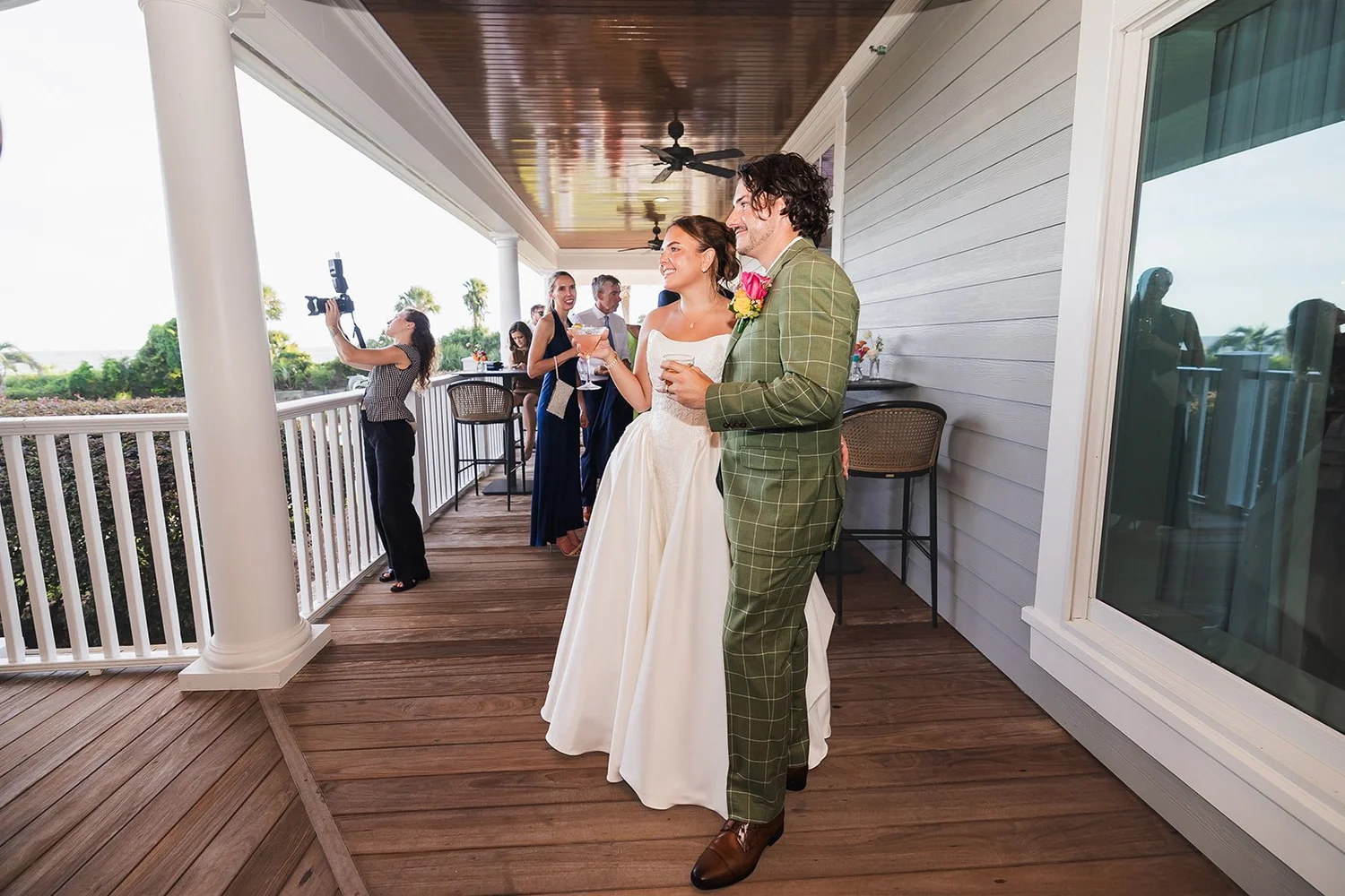 Karolina and Connor hold their drinks on a covered clubhouse veranda with white railings as guests gather behind them at Seabrook Island Club on Seabrook Island, South Carolina.