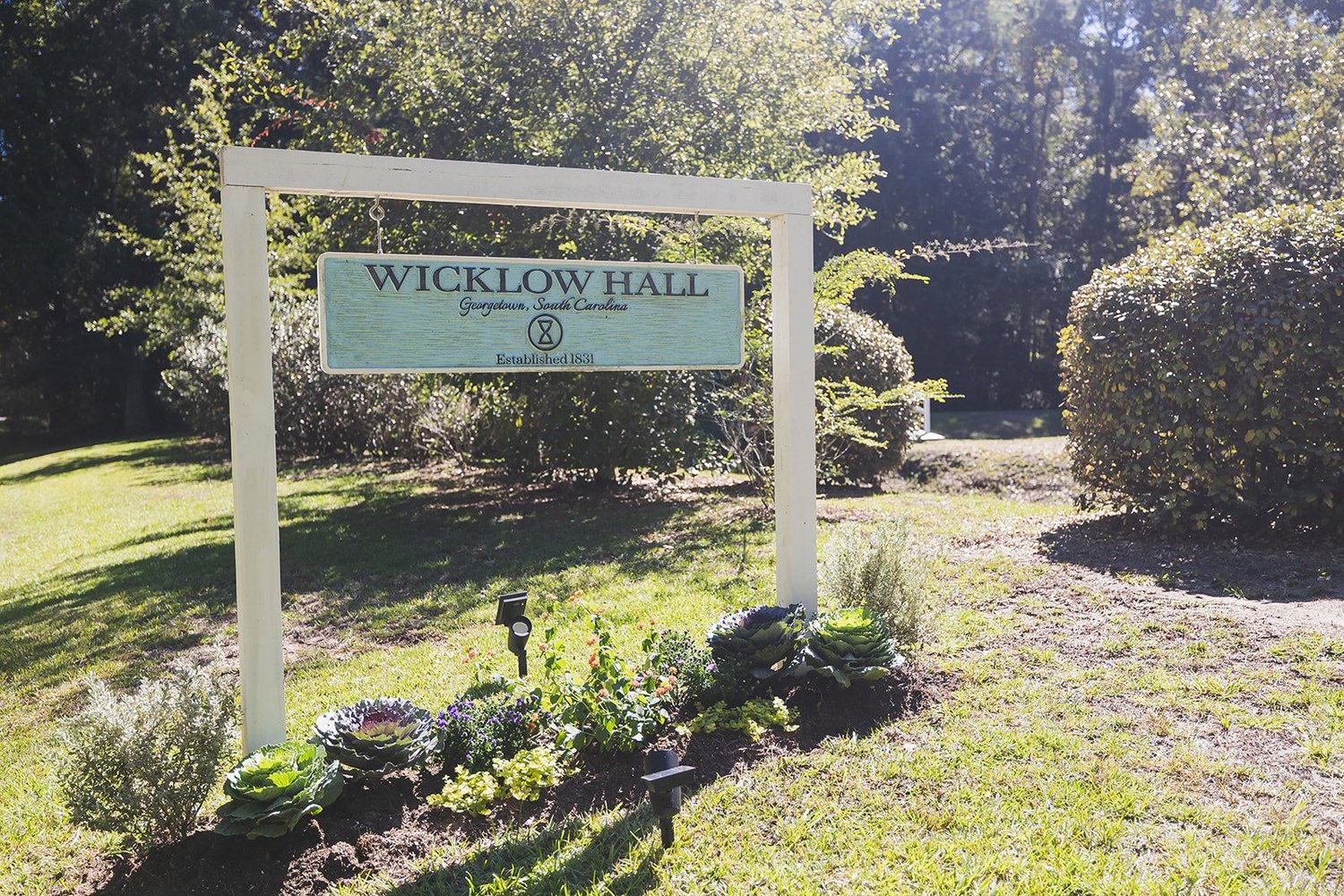 The white wooden entrance sign for Wicklow Hall in Georgetown, South Carolina, with gardens planted below it and trees behind.