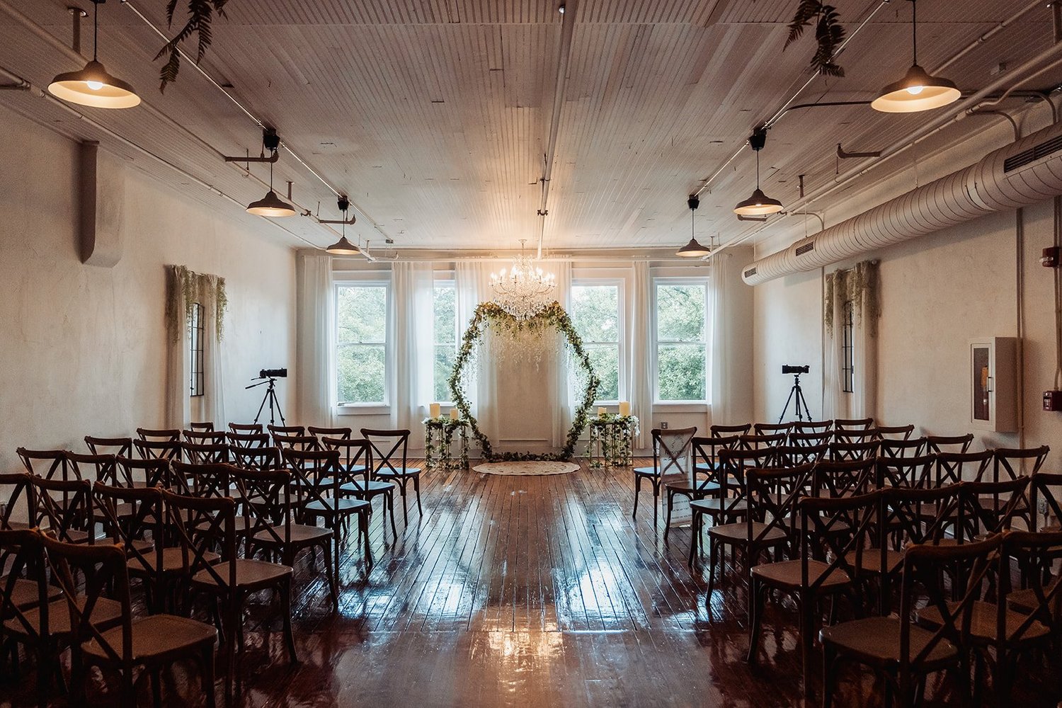 An indoor wedding ceremony set at Vici Rooftop in Savannah, Georgia, with rows of wooden cross-back chairs, a greenery arch beneath a crystal chandelier, tall windows and glossy wood floors.