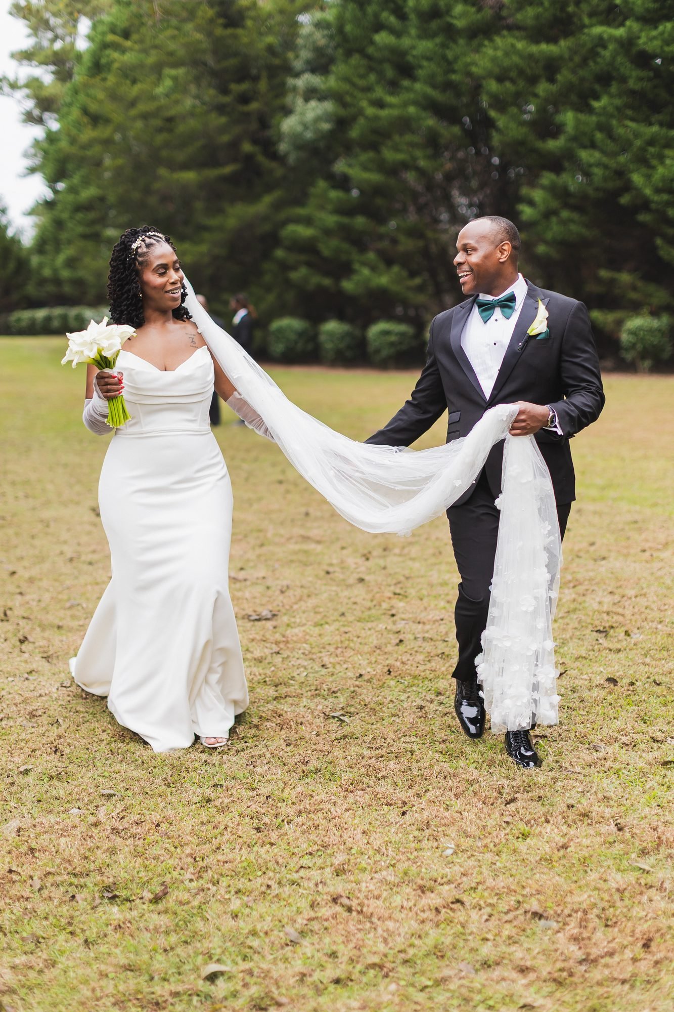 Ashlee and Chris wedding at Southern Oaks Event Venue in Gilbert, South Carolina: couple walking on the lawn as the groom holds the bride's long veil.