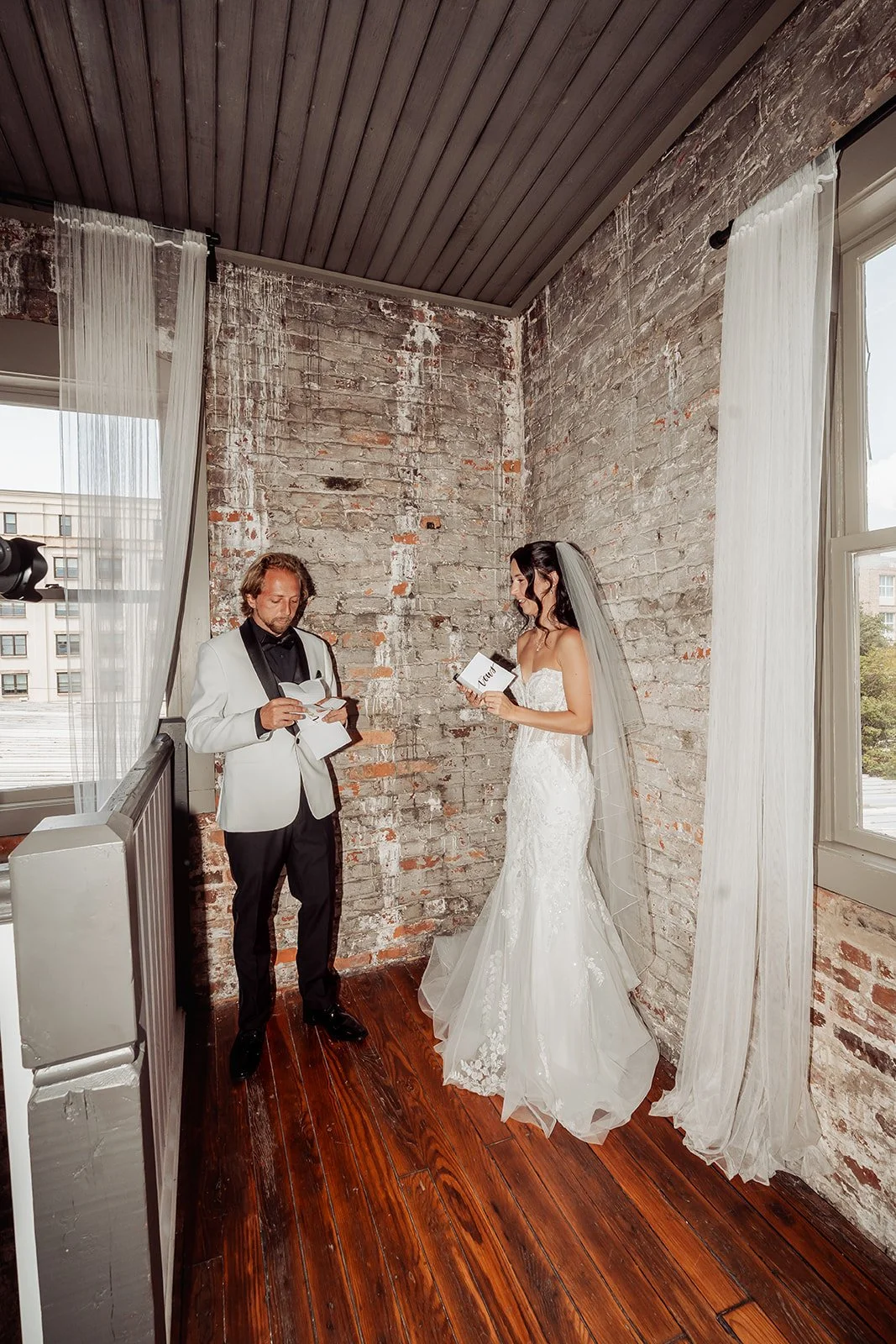 Brienna and Michael read their vows to each other in a corner of exposed brick and tall windows, with downtown Savannah buildings outside, at Vici Rooftop in Savannah, Georgia.