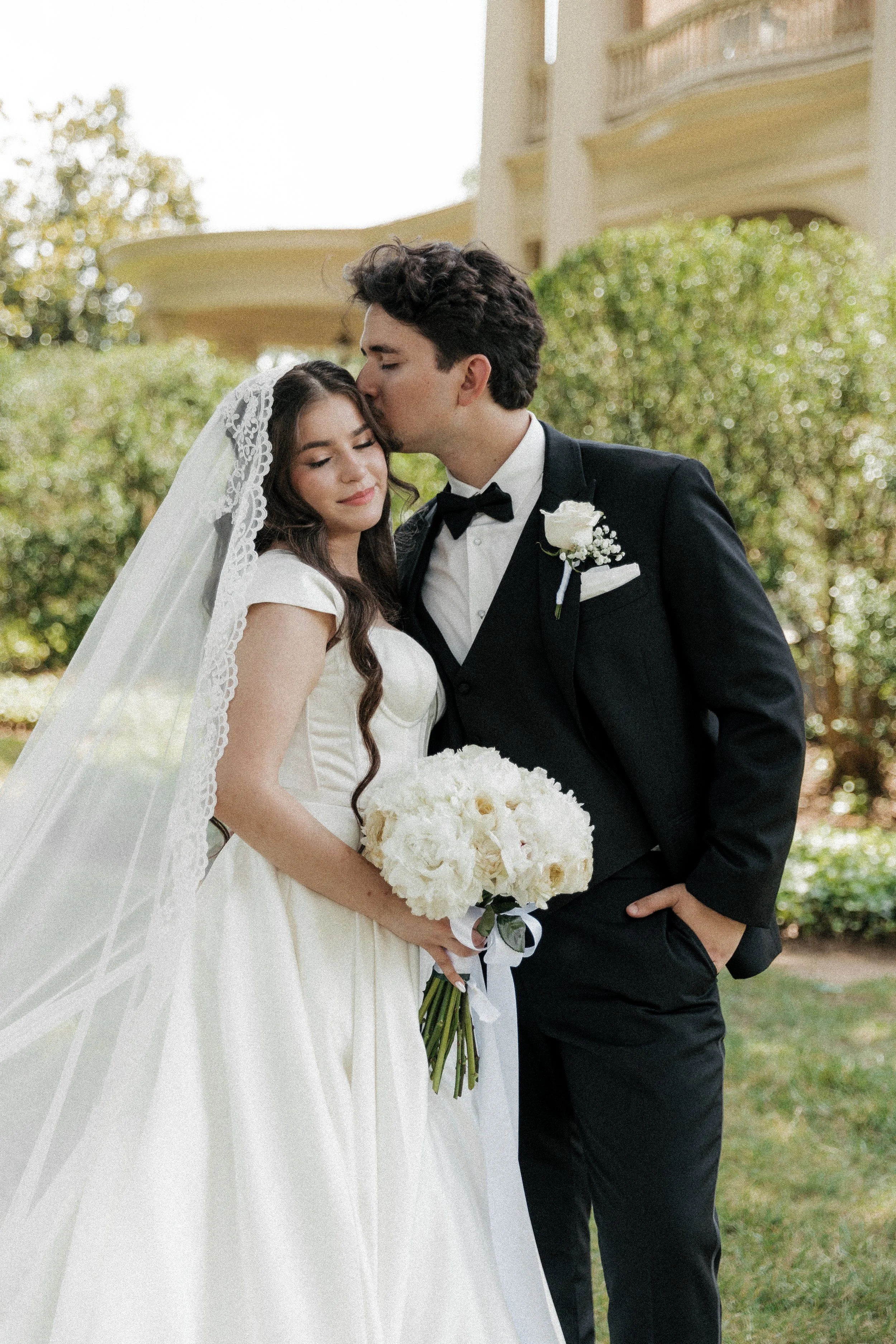 A bride and groom are close together outdoors; the groom is kissing the bride's forehead, and she is holding a bouquet of white flowers.