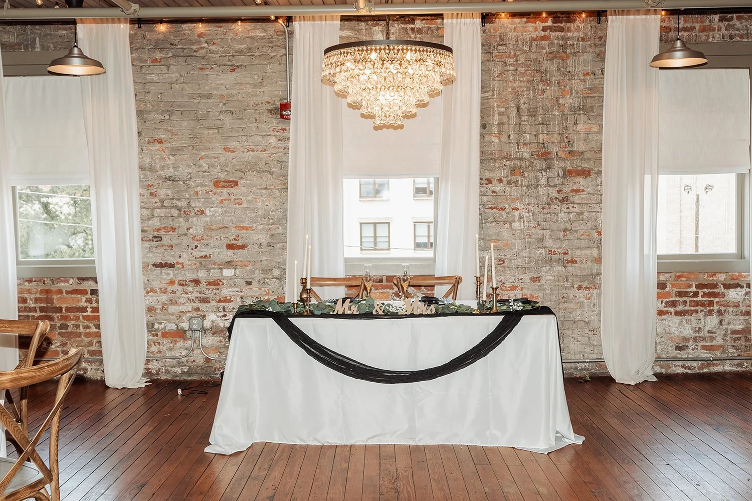 Brienna and Michael's sweetheart table against exposed brick, beneath a crystal chandelier and between tall draped windows, set indoors at Vici Rooftop in Savannah, Georgia.