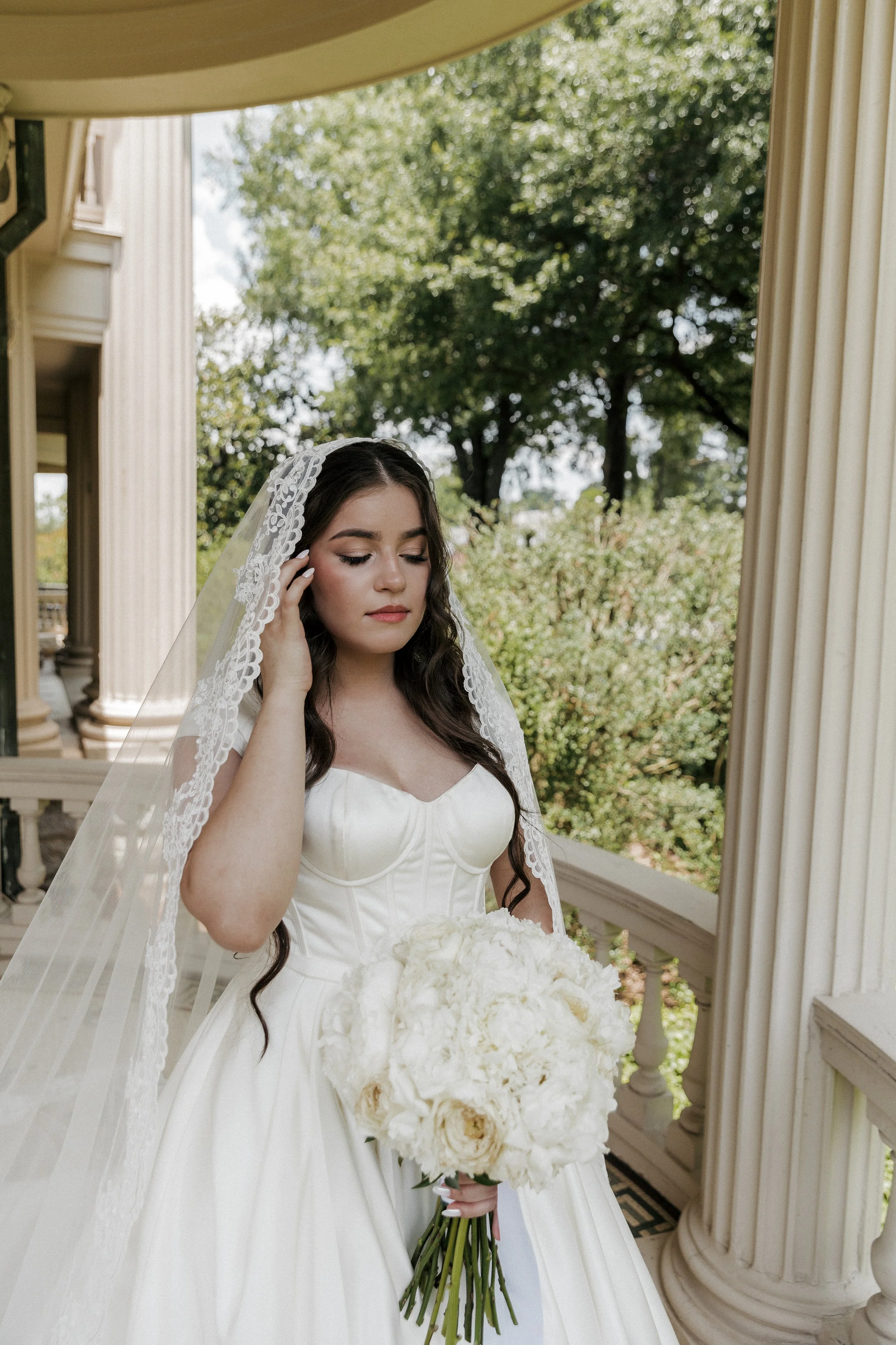 A bride in a white wedding dress with a veil holds a bouquet of white flowers, standing outdoors next to white columns and a garden with trees.