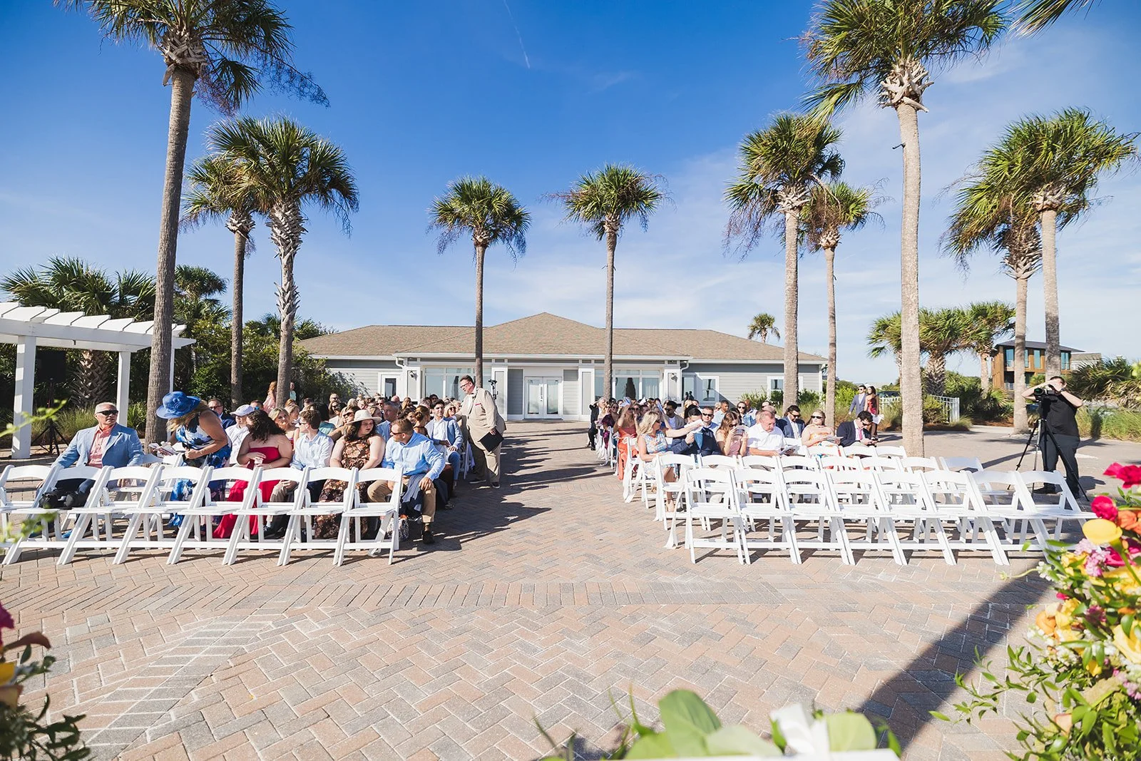Guests seated in rows of white chairs on a brick terrace lined with tall palms, with the clubhouse and a white pergola behind them, at Karolina and Connor's ceremony at Seabrook Island Club on Seabrook Island, South Carolina.