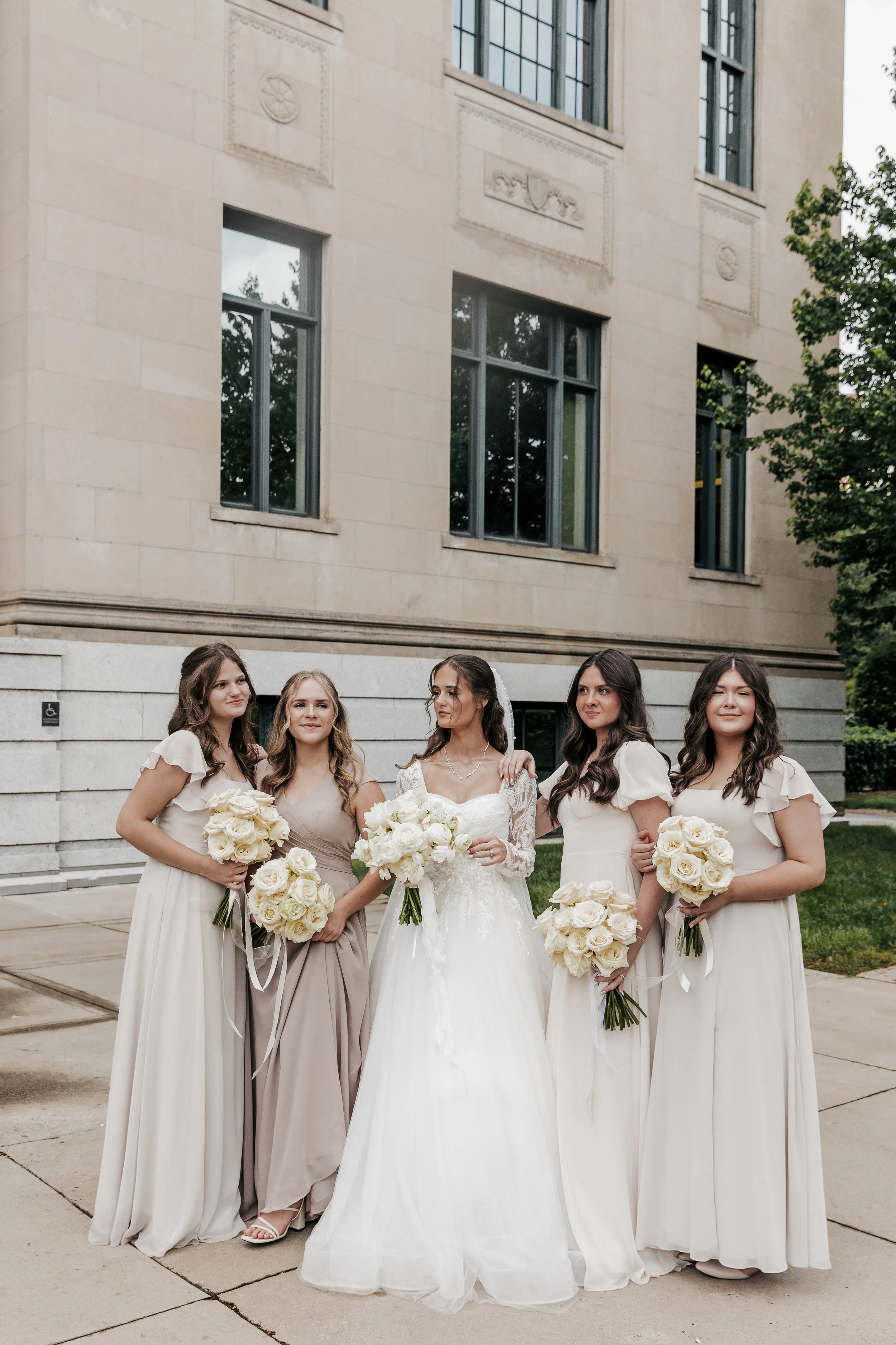 A bride in a white wedding gown with four bridesmaids in light-colored dresses holding bouquets of white roses outside a building with large windows and stone facade.