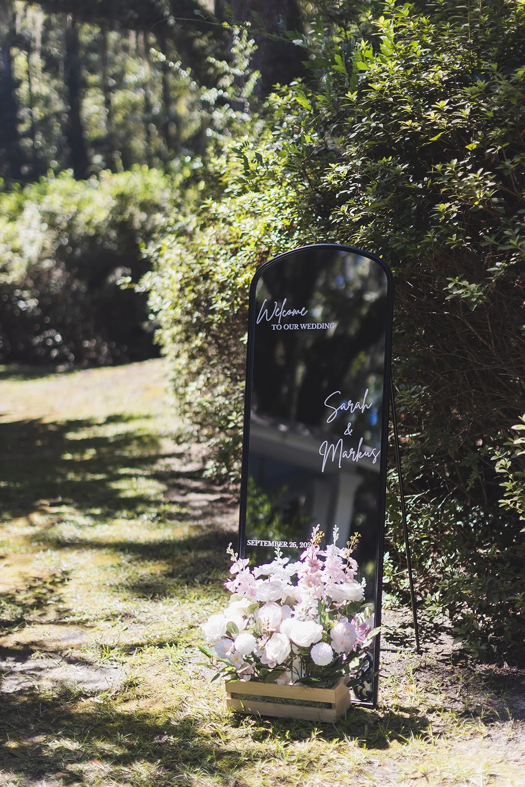 A black arched welcome mirror reading Sarah and Markus, with blush florals at its base, on the grounds at Wicklow Hall in Georgetown, South Carolina.