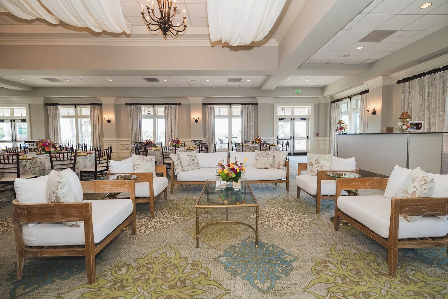 A clubhouse lounge with draped ceilings, a chandelier, white sofas and French doors, with dining tables set beyond, at Seabrook Island Club on Seabrook Island, South Carolina.