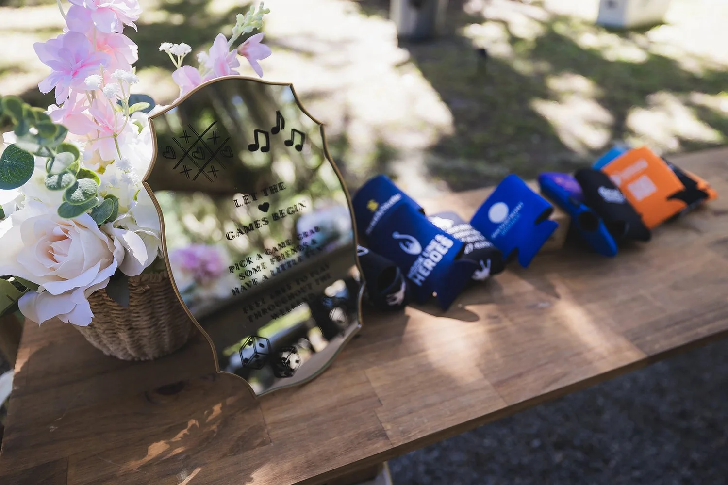 Custom koozies lined up on a wooden table beside a mirror sign and pink florals for guests at Wicklow Hall in Georgetown, South Carolina.