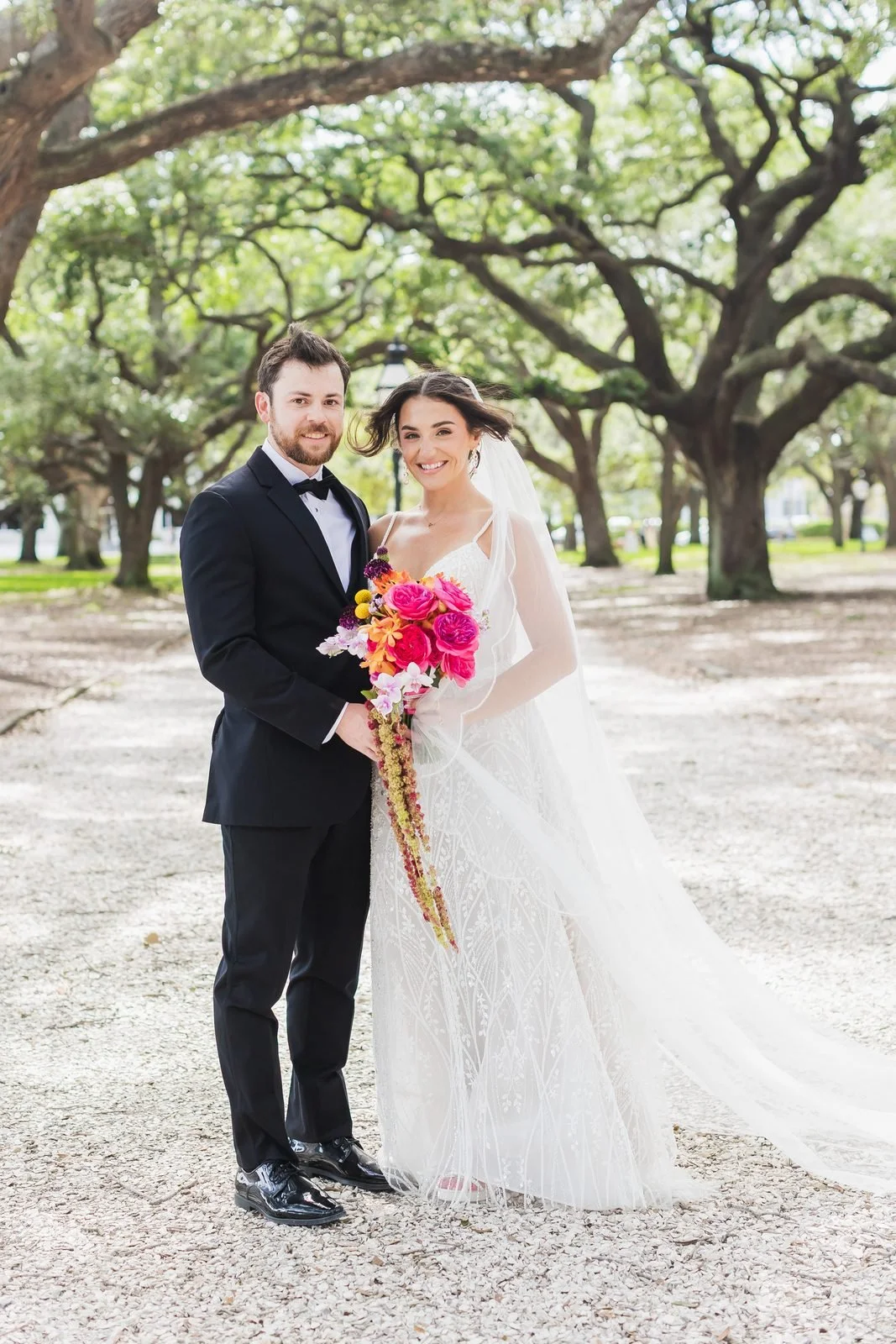 Full-length bridal portrait under live oaks with a windblown veil and bright pink and orange bouquet, Charleston area
