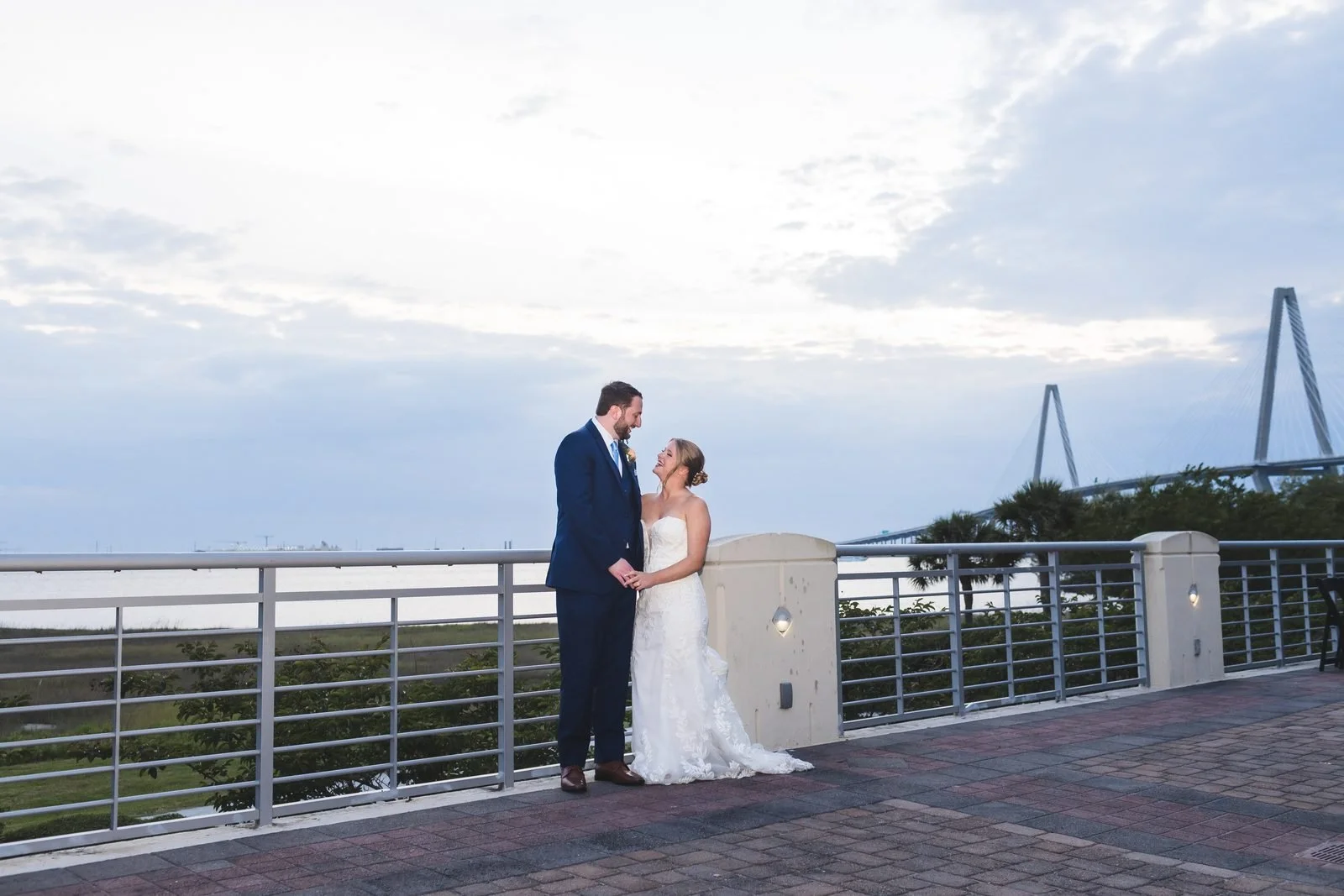 Bride and groom on a waterfront terrace at blue hour under a big sky, the Ravenel Bridge behind them in Charleston