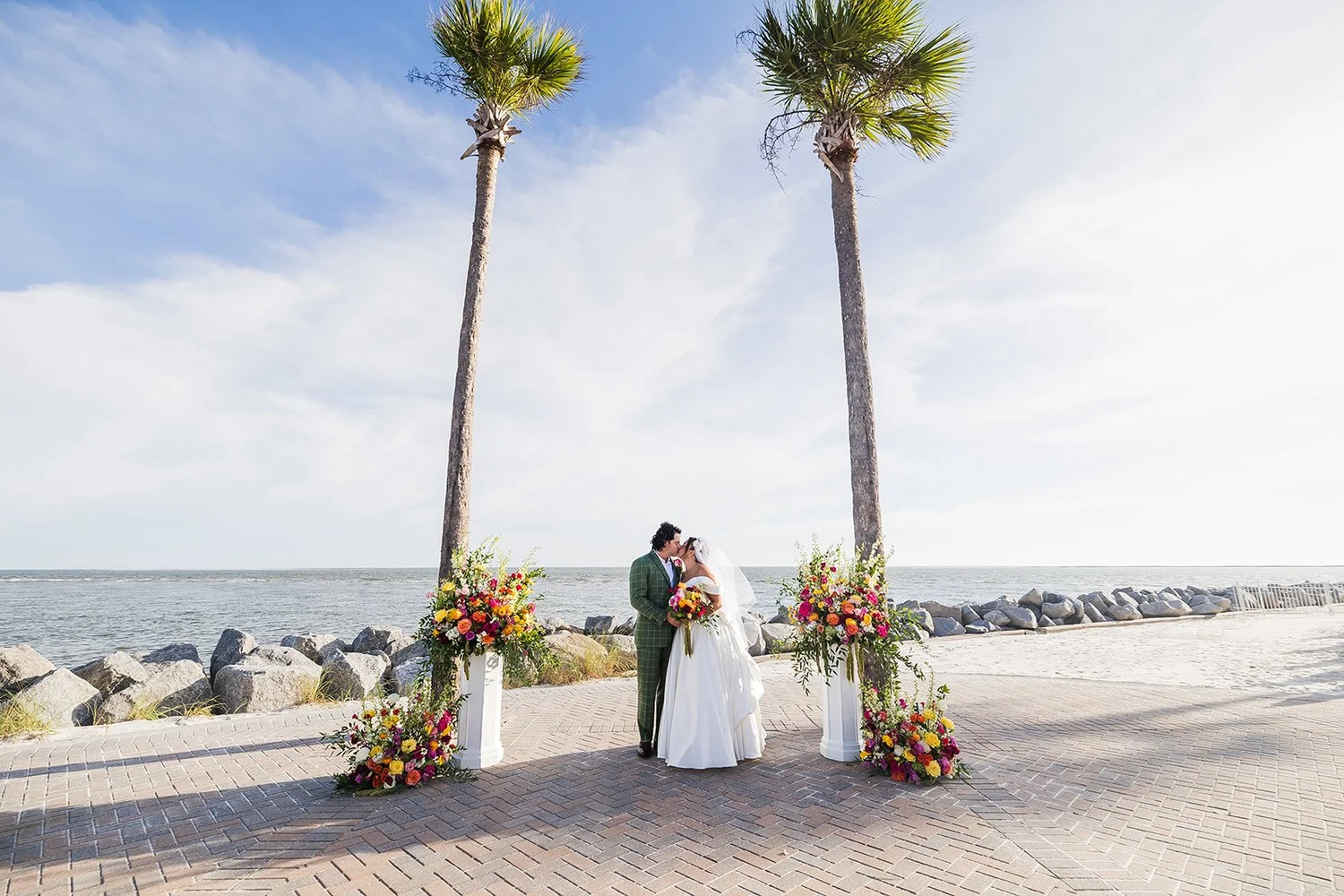 Karolina and Connor stand between two tall palms and bright floral arrangements on white pedestals, with the rocky seawall and the ocean behind them, at Seabrook Island Club on Seabrook Island, South Carolina.