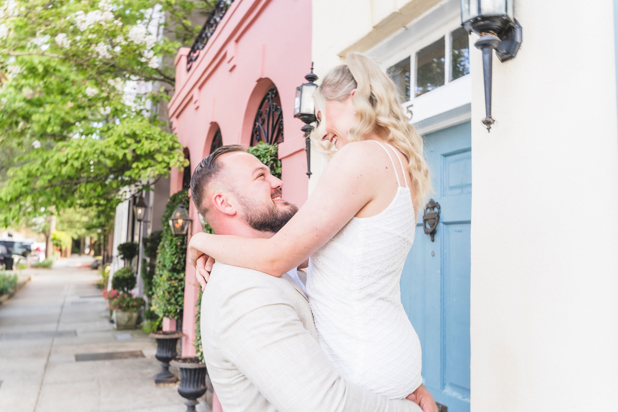 Groom lifting fiancée in front of pastel houses on Rainbow Row, Charleston