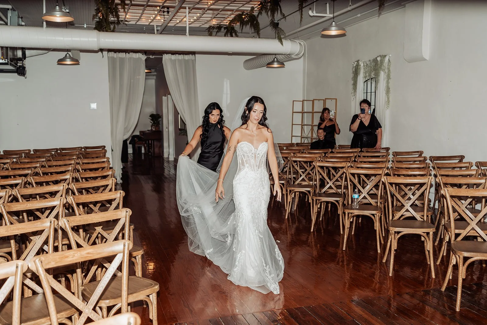 Brienna walks down the aisle as her bridesmaid carries her veil, at her indoor wedding ceremony at Vici Rooftop in Savannah, Georgia.