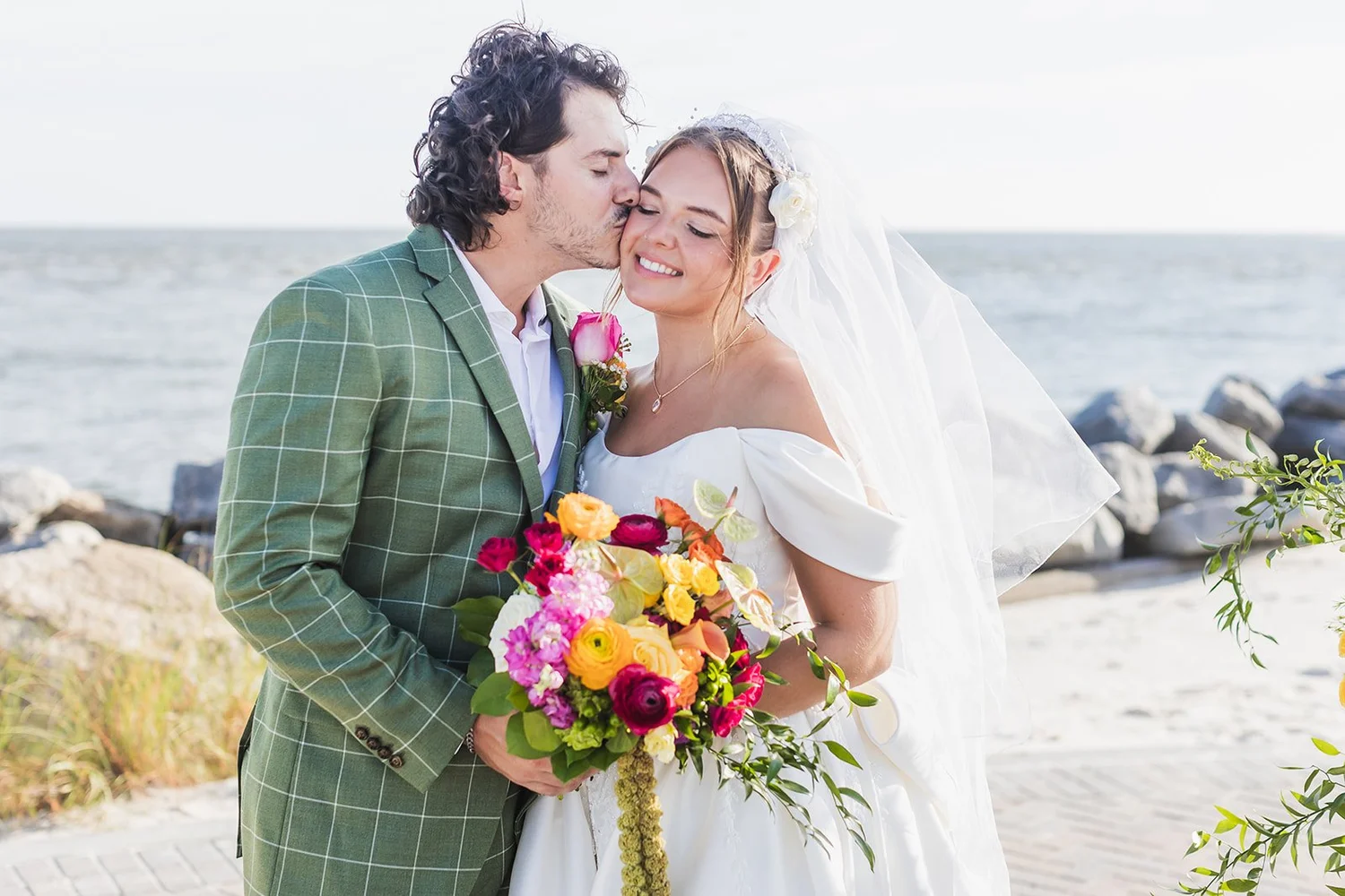 Connor, in a green plaid suit, kisses Karolina on the cheek as she holds her bright bouquet, with the seawall and ocean behind them, at Seabrook Island Club on Seabrook Island, South Carolina.