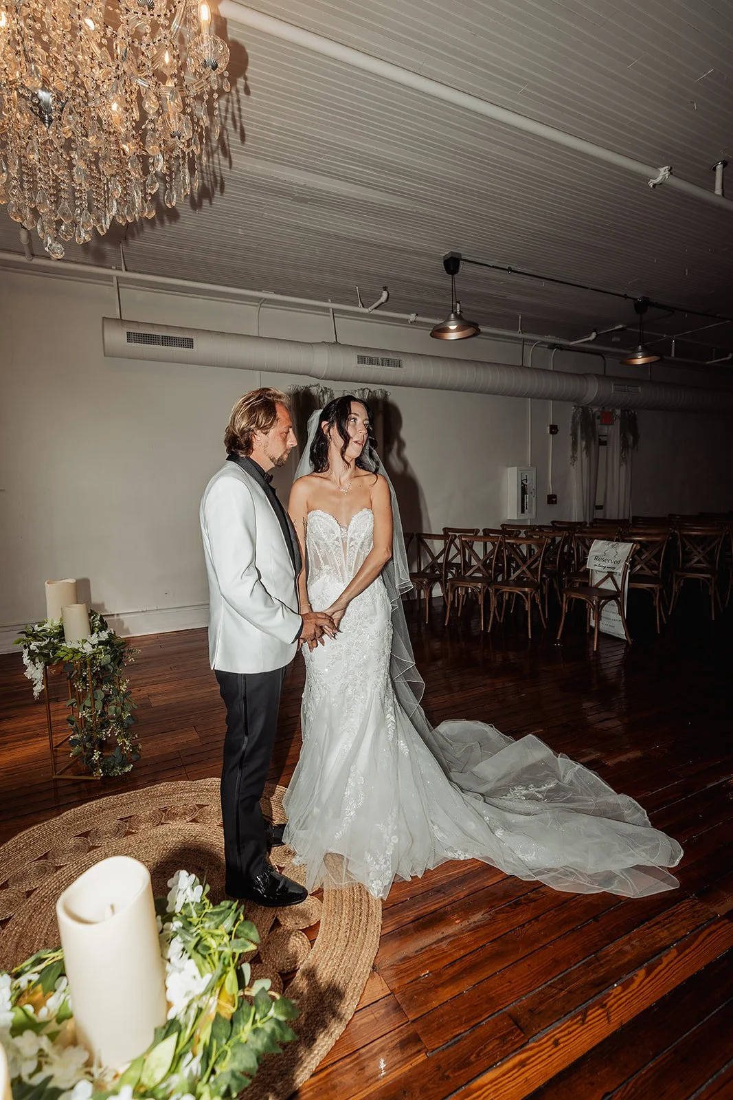 Brienna and Michael stand together beneath a crystal chandelier in the indoor ceremony room at Vici Rooftop in Savannah, Georgia.
