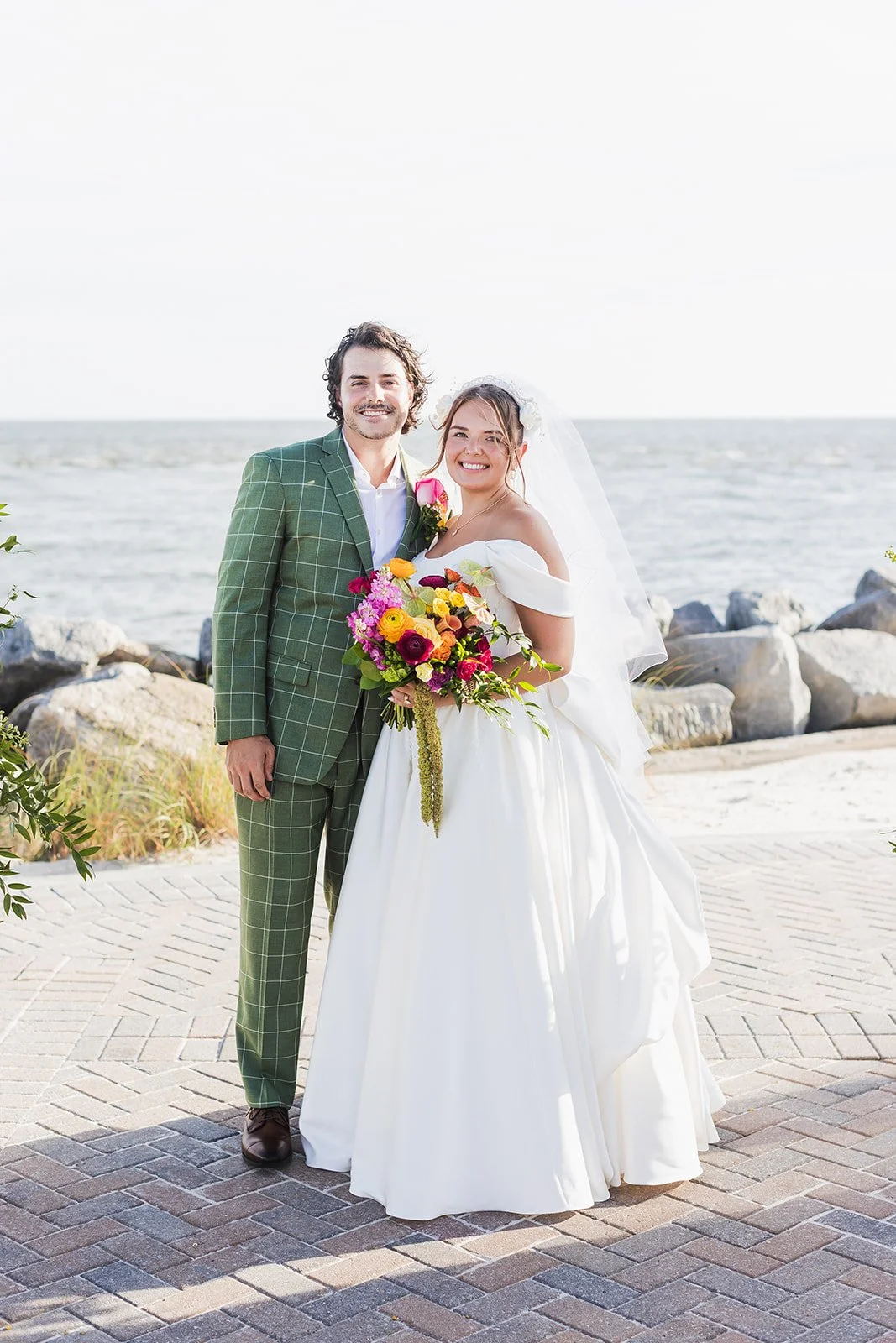 Karolina and Connor smile together on a brick terrace with the rocky seawall and the ocean behind them at Seabrook Island Club on Seabrook Island, South Carolina.