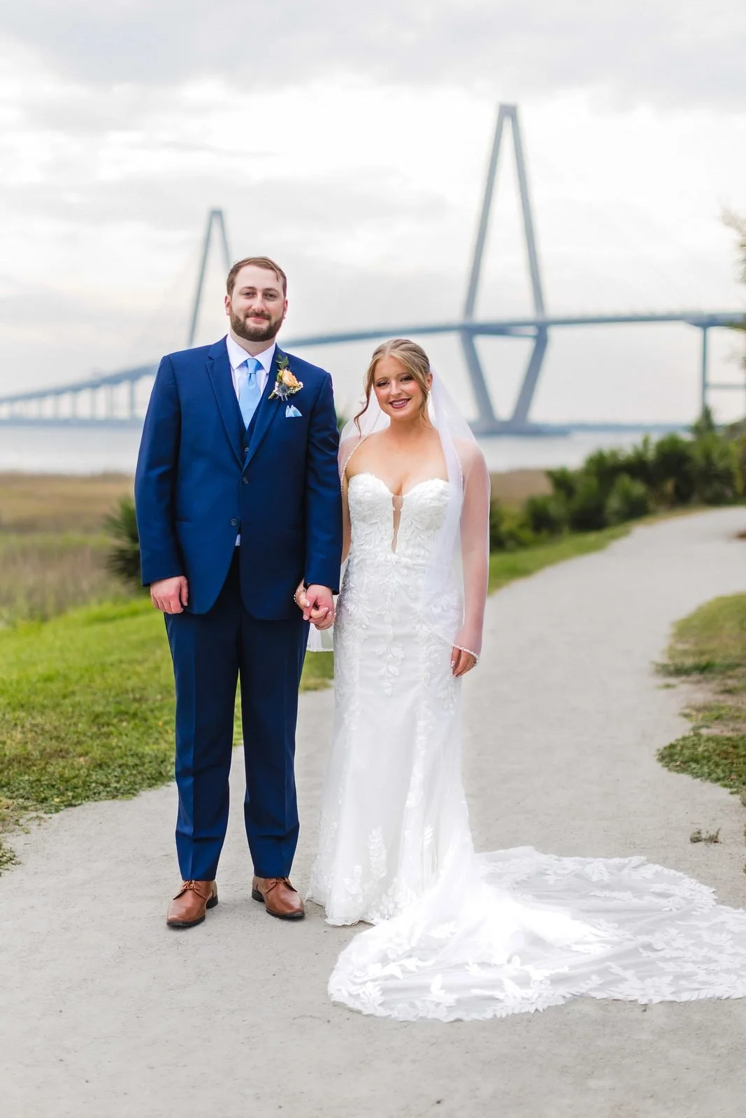 Full-length portrait of a bride with a long lace train and her groom, a Ravenel Bridge tower behind them in Charleston