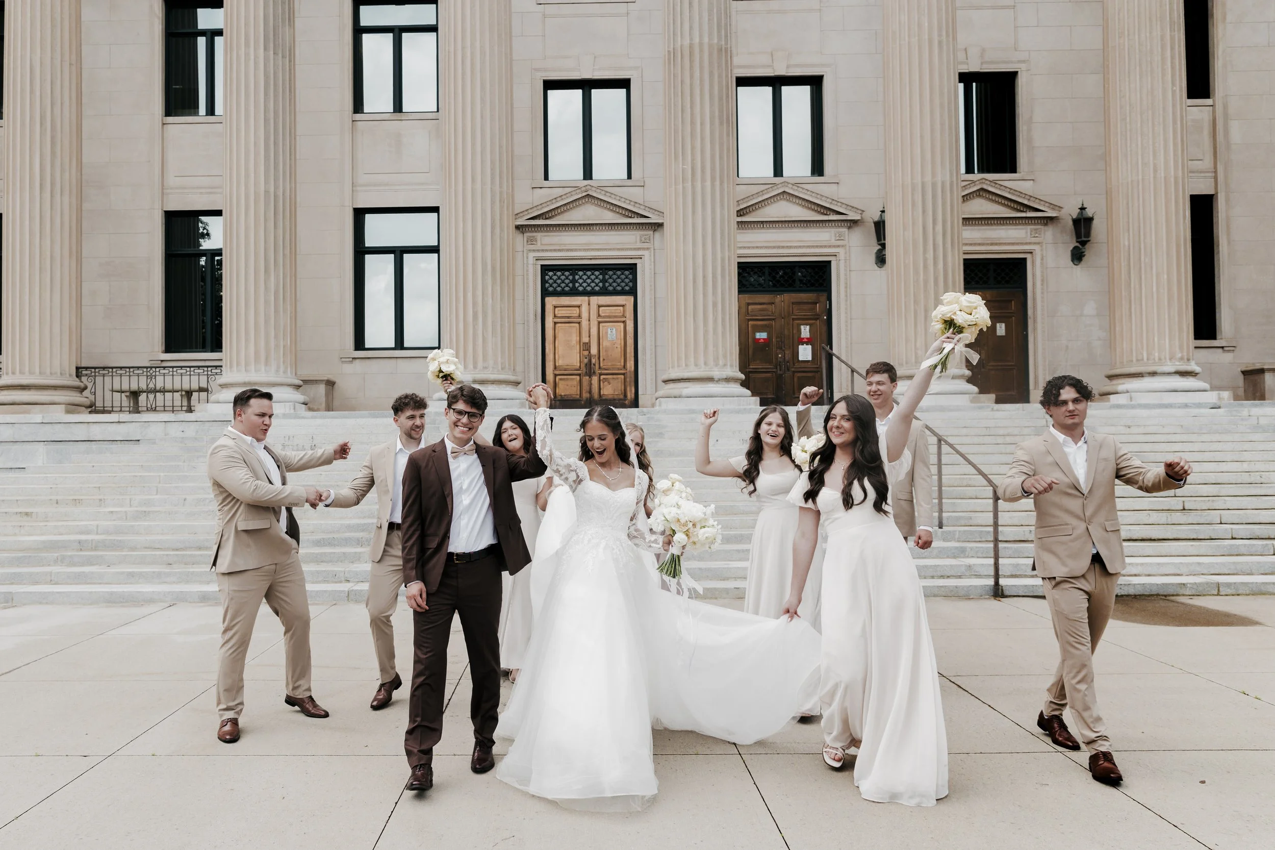 A wedding celebration outside a grand building with large columns. The bride, in a white wedding dress, is holding a bouquet and walking with her bridal party, who are dressed in cream or white and brown suits. The group is smiling, cheering, and cel