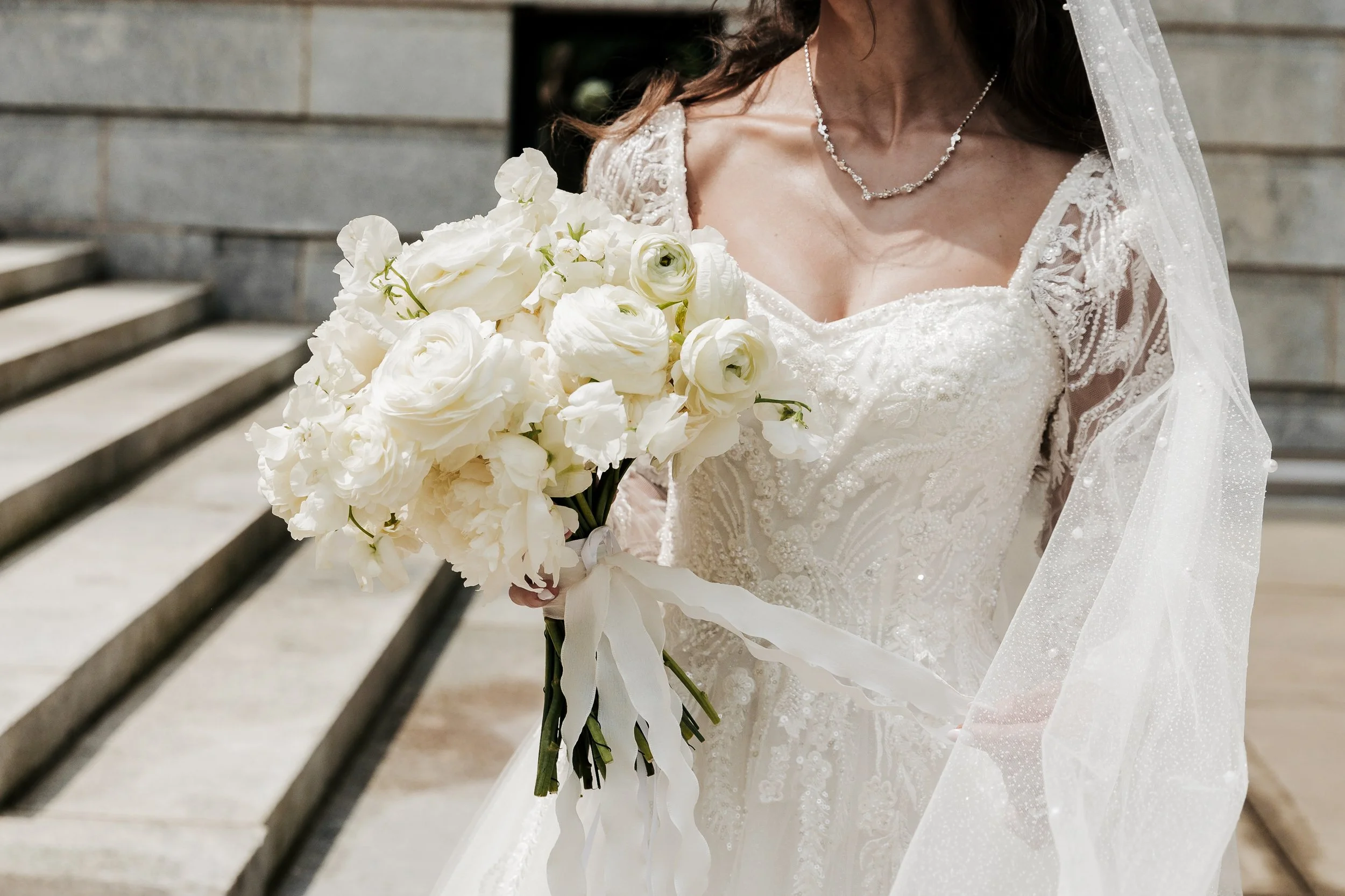 Bride in a white wedding dress holding a bouquet of white flowers near a stone staircase.