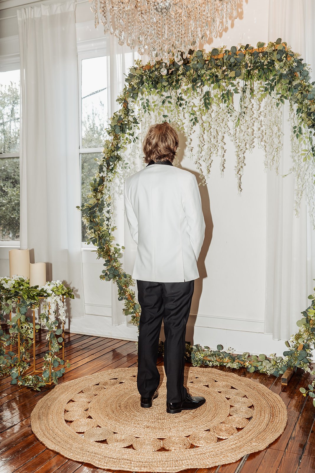 Michael stands on a round jute rug facing the greenery arch beneath a crystal chandelier, indoors at Vici Rooftop in Savannah, Georgia.
