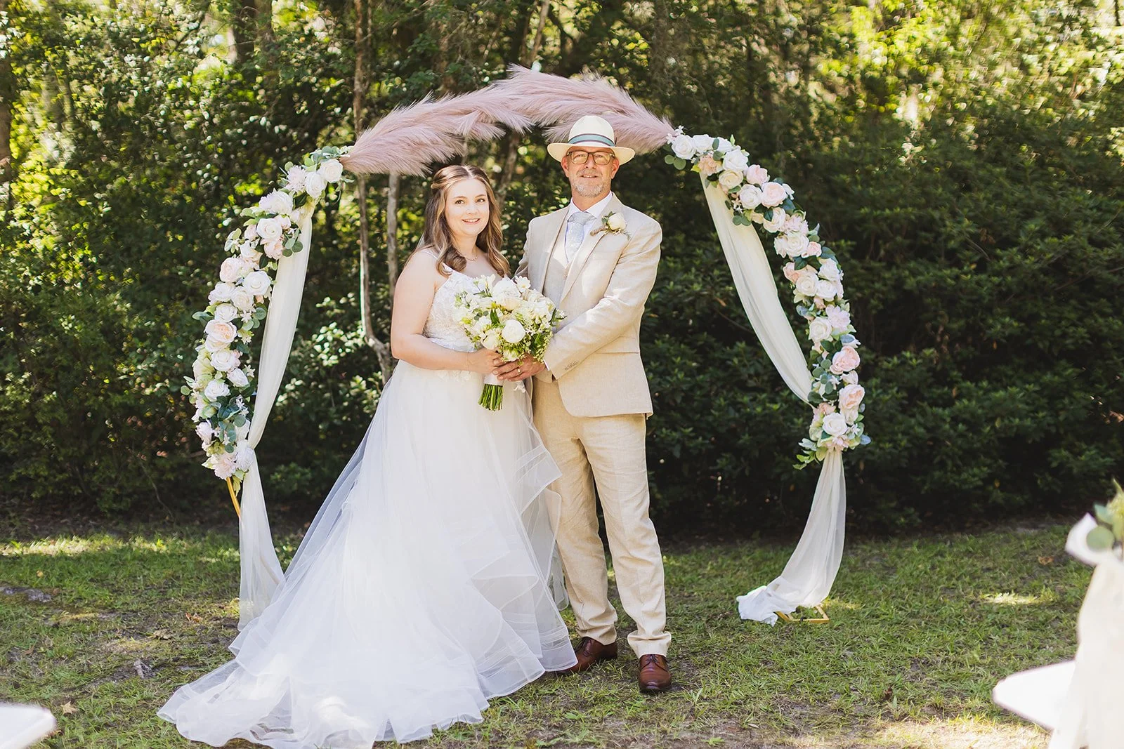 Sarah and Markus smile in front of a white floral arch topped with pink pampas grass on the lawn at Wicklow Hall in Georgetown, South Carolina.