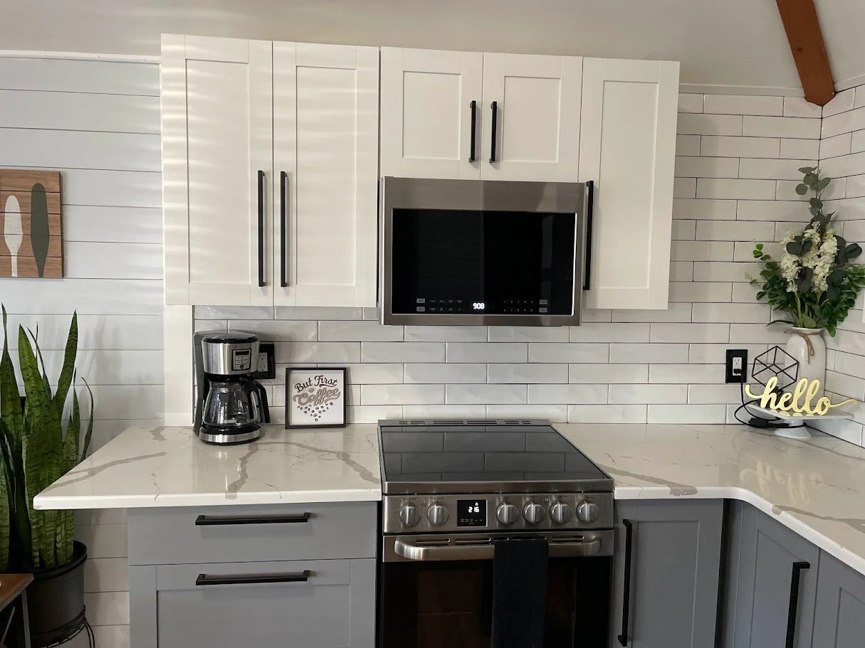 Modern kitchen with white cabinets, stainless steel microwave above stove, coffee maker, decorative sign, flower arrangement, and potted plant on countertop.