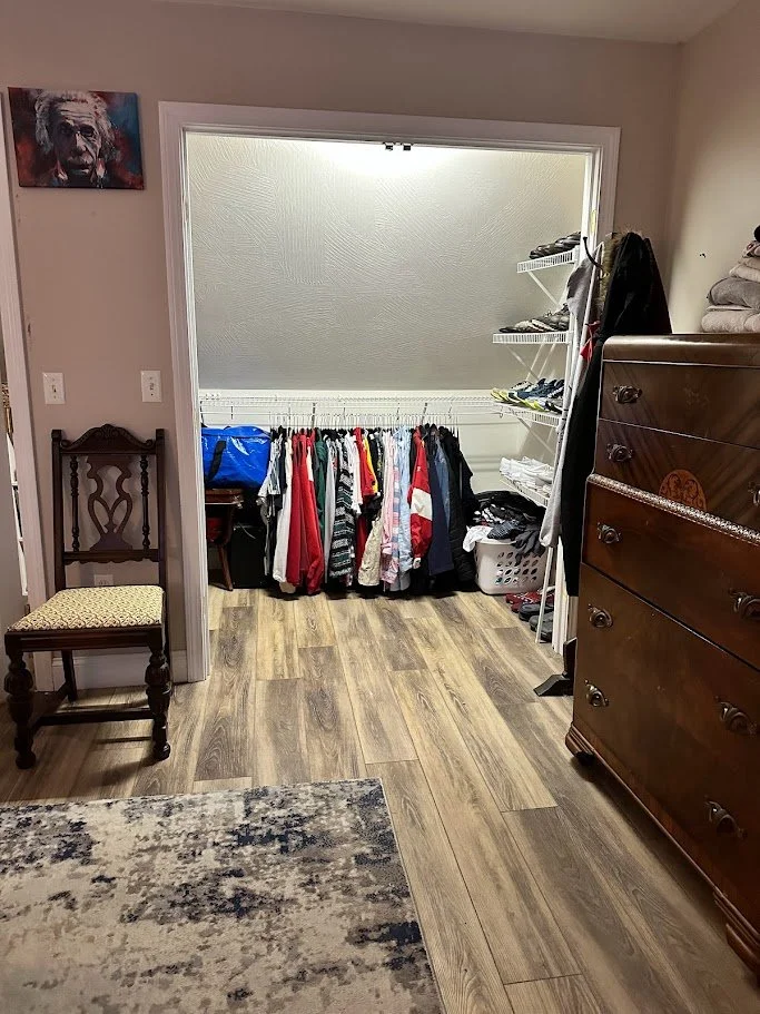 A closet with hanging clothes, shelves with shoes, and a laundry basket, next to a dark wooden dresser and a decorative wooden chair with a patterned cushion.
