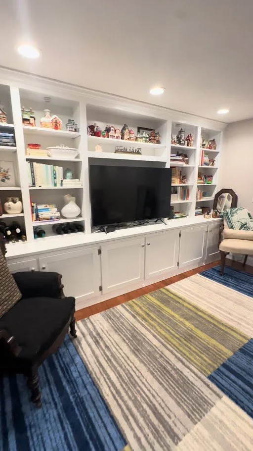 Living room with white built-in bookshelves filled with books and decorative items, a large flat-screen TV, a black armchair, a beige armchair with a patterned pillow, and a colorful striped rug on a hardwood floor.