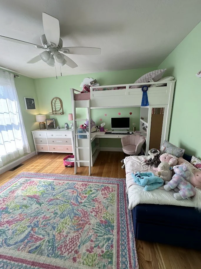 A child's bedroom with mint green walls, a mold white loft bed with pink bedding, a desk with a computer and plush toys underneath, a pink chair, a floral rug, a white dresser with lamps and picture frames, and a navy sofa with stuffed animals.