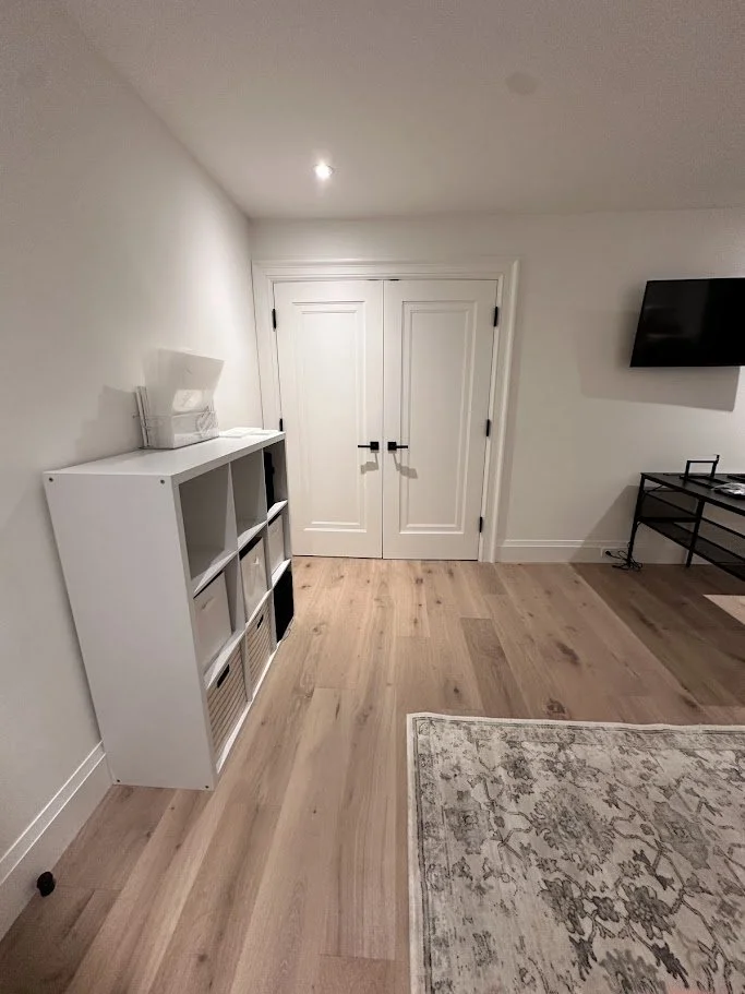 Room with white double doors, light wood flooring, a white bookshelf on the left, and a flat-screen TV on the right wall.