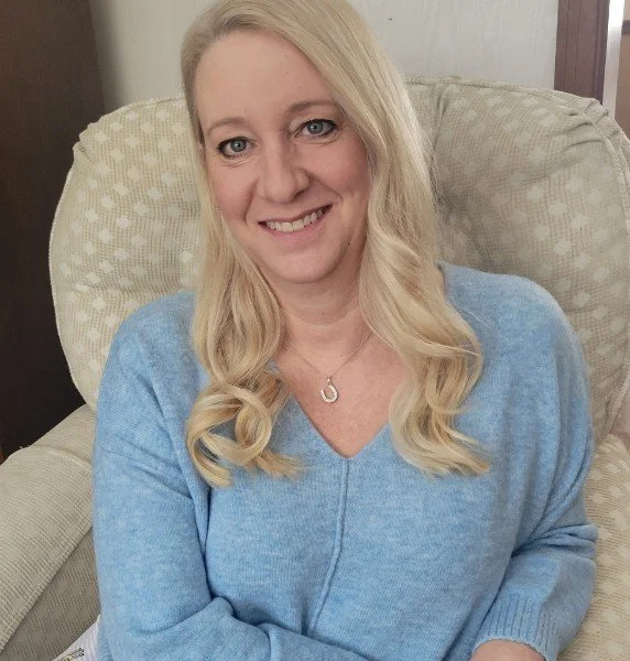 Woman with long blonde hair in a blue sweater sitting on a beige chair, smiling.