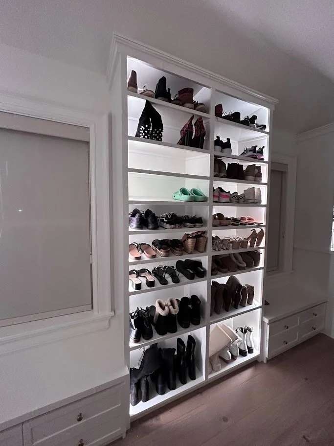 A white shoe shelf with multiple rows of various shoes and boots, illuminated by integrated lighting, in a room with white walls and wooden flooring.