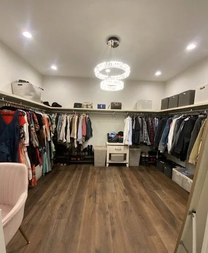 A walk-in closet with clothing on racks, storage boxes on shelves, a chandelier, and a chair along the wall.