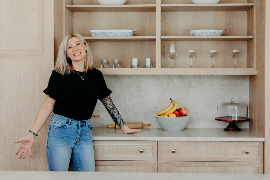 Smiling person in kitchen with wooden cabinets, open shelves, bowls, glasses, and a fruit bowl.