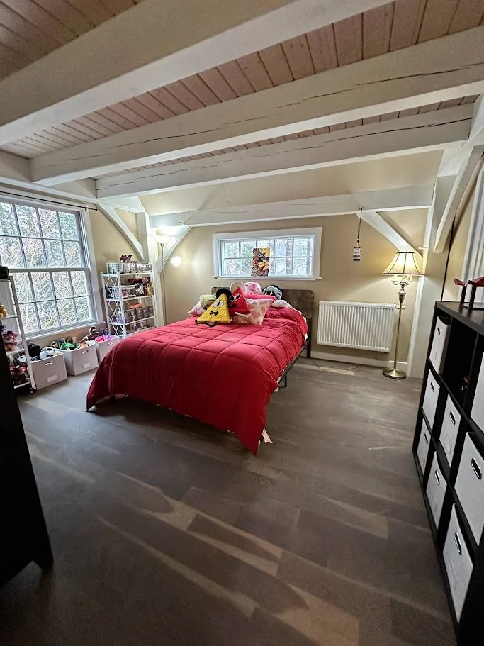 A cozy bedroom with a sloped ceiling and wooden beams, featuring a bed with a red comforter and various pillows, a white shelving unit filled with toys, a large window, a small window above the bed, and a floor lamp.